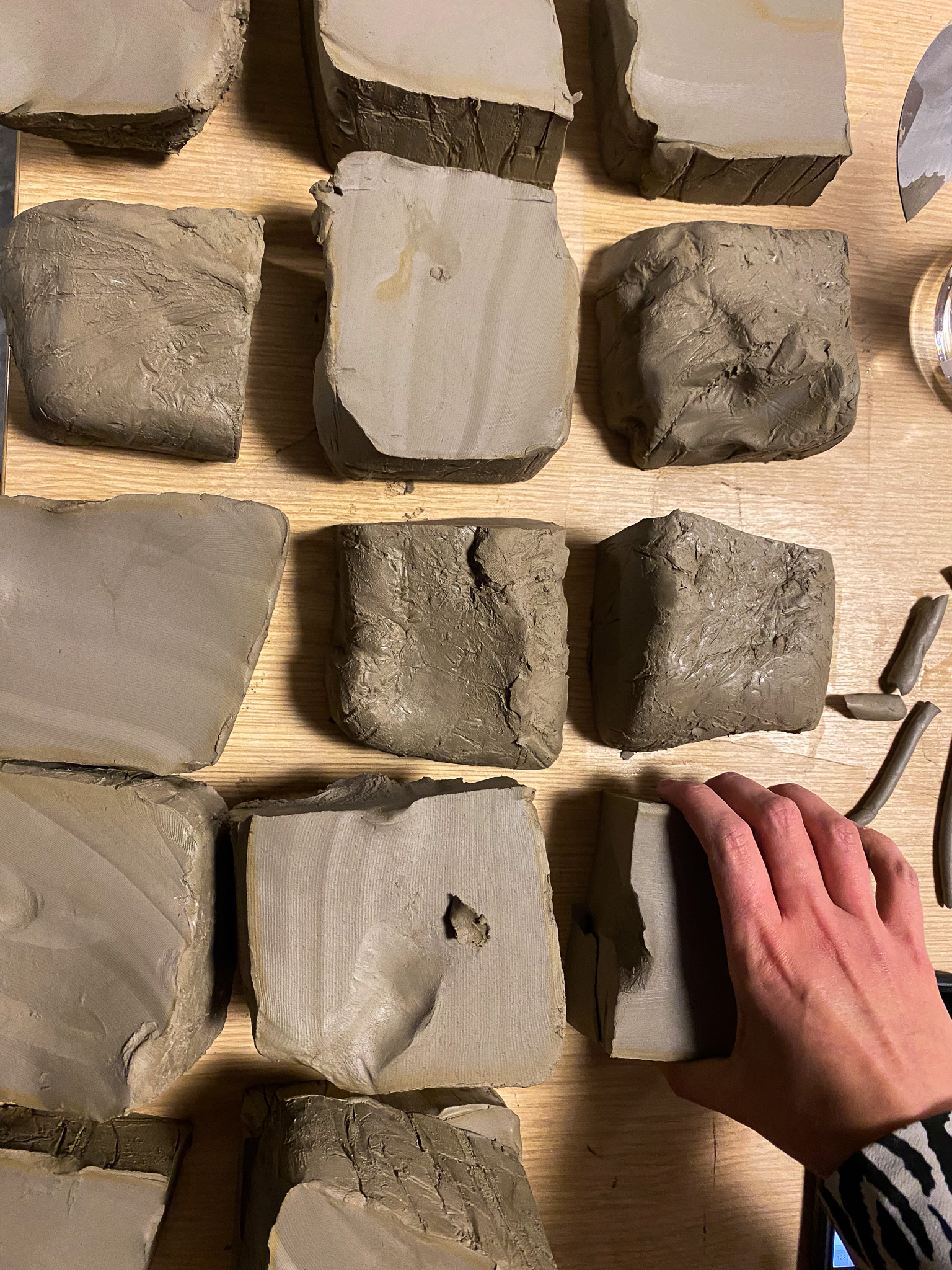 Several rectangular blocks of clay are arranged on a wooden surface. A hand is touching one of the blocks in the lower right corner, and sculpting tools are partially visible nearby.