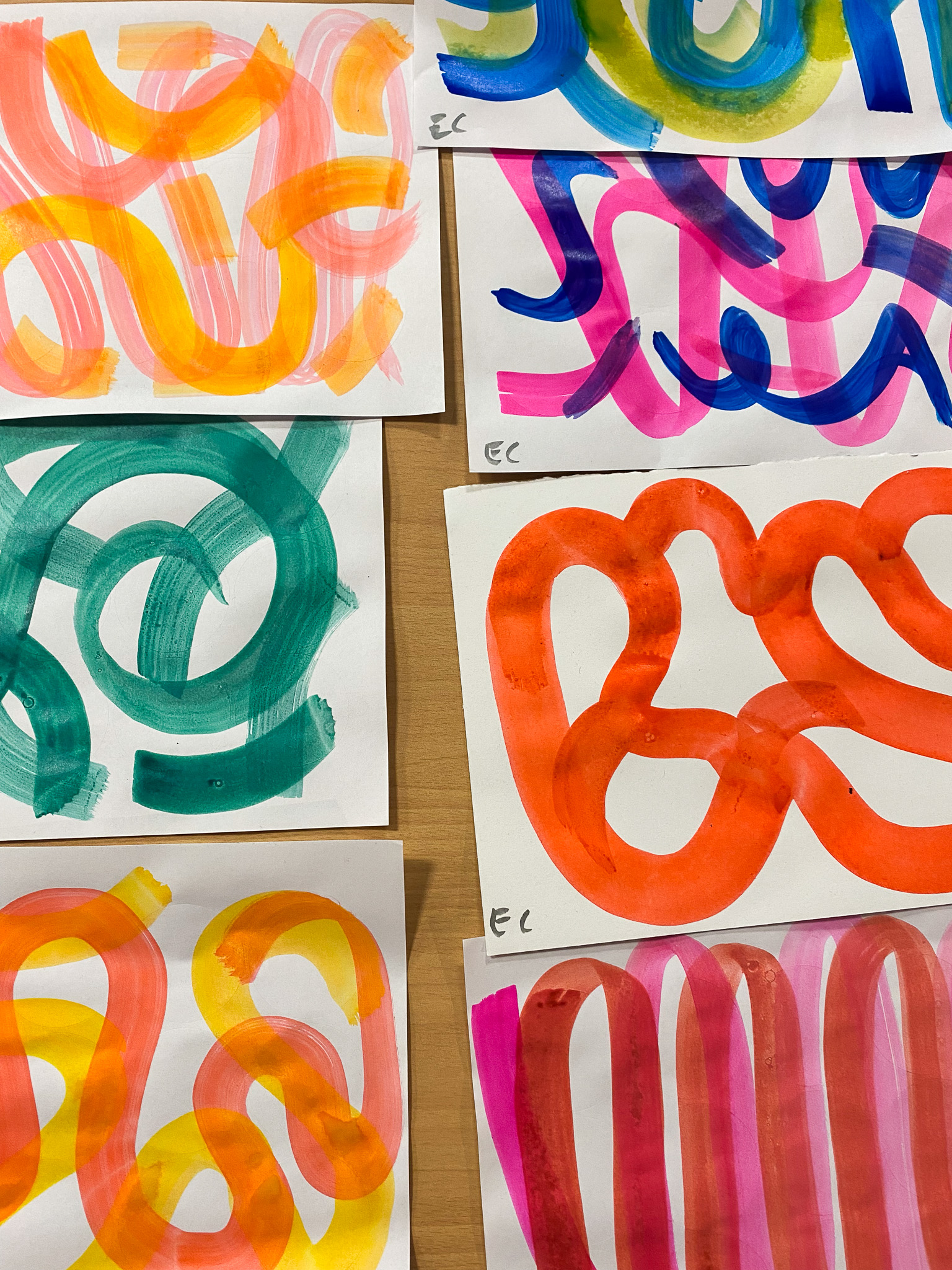 Several abstract paintings on paper are displayed on a wooden surface. Each painting features colourful, curving, and intertwining lines in various bright colours like orange, green, pink, yellow, and blue.