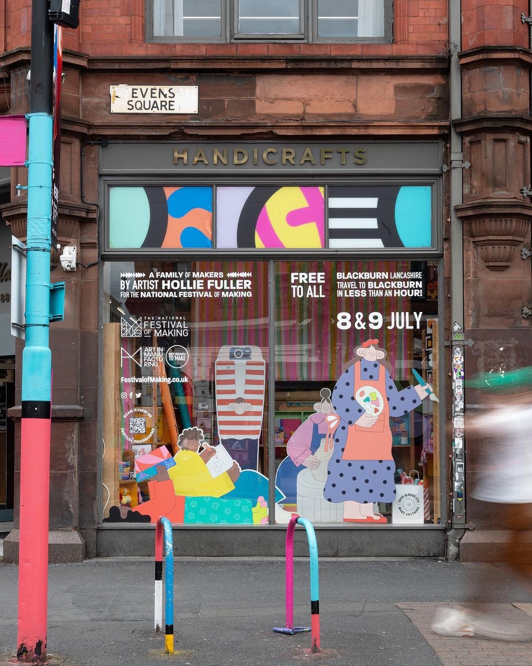 A shopfront with a sign reading HANDICRAFTS displays colourful artwork and text promoting a free festival by artist Hollie Fuller. The window is decorated with illustrated characters and event details for 8 & 9 July.