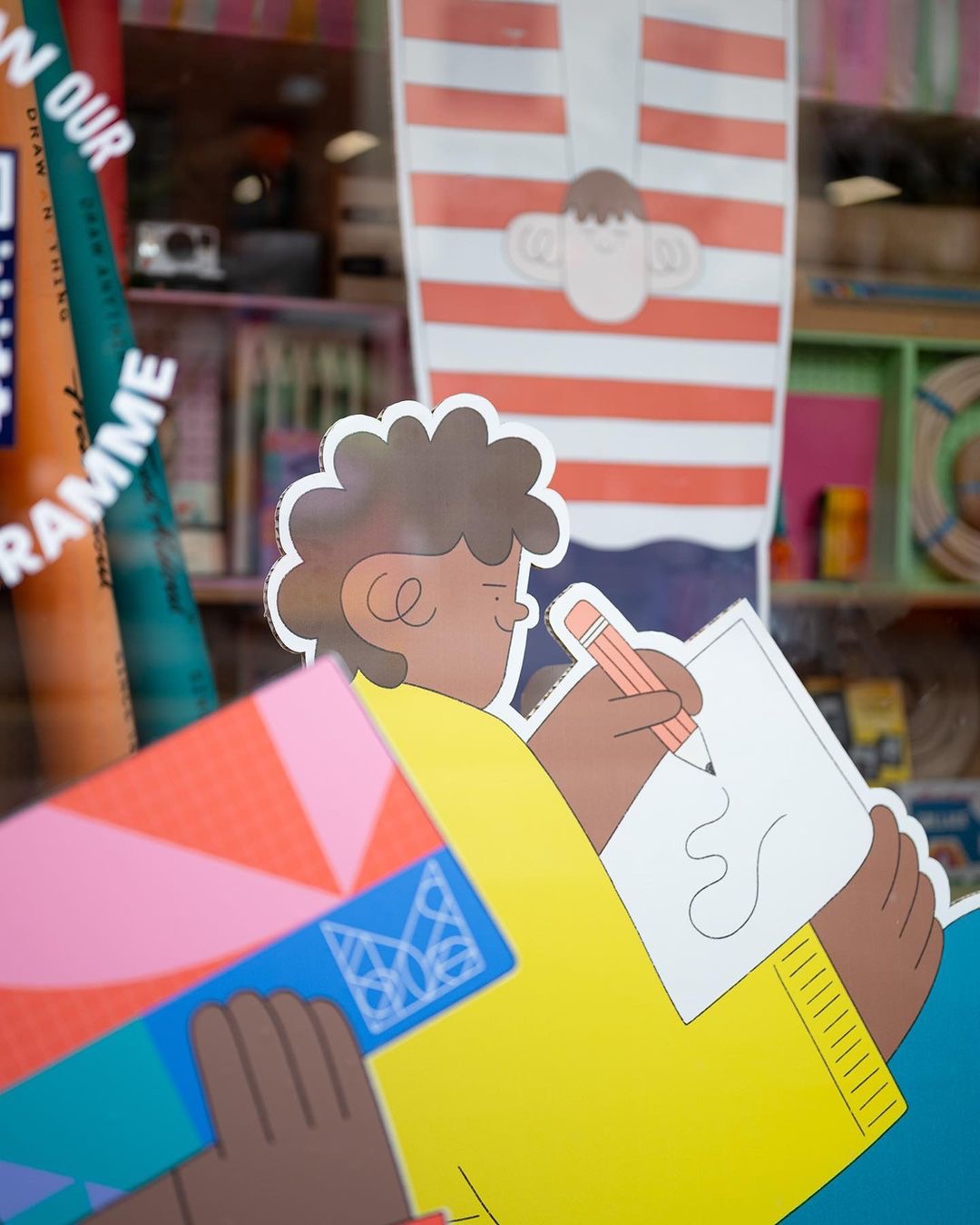 A colourful window display features a cartoon illustration of a child with curly hair drawing on paper, holding a large pink crayon. Another cartoon character in a red-striped shirt appears upside down above them.