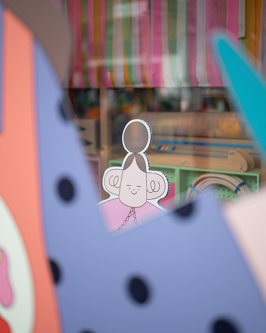 A paper cut-out of a smiling character with a bun hairstyle is displayed behind colourful, abstract shapes and a striped background. The scene appears playful and artistic, with soft focus and pastel colours.