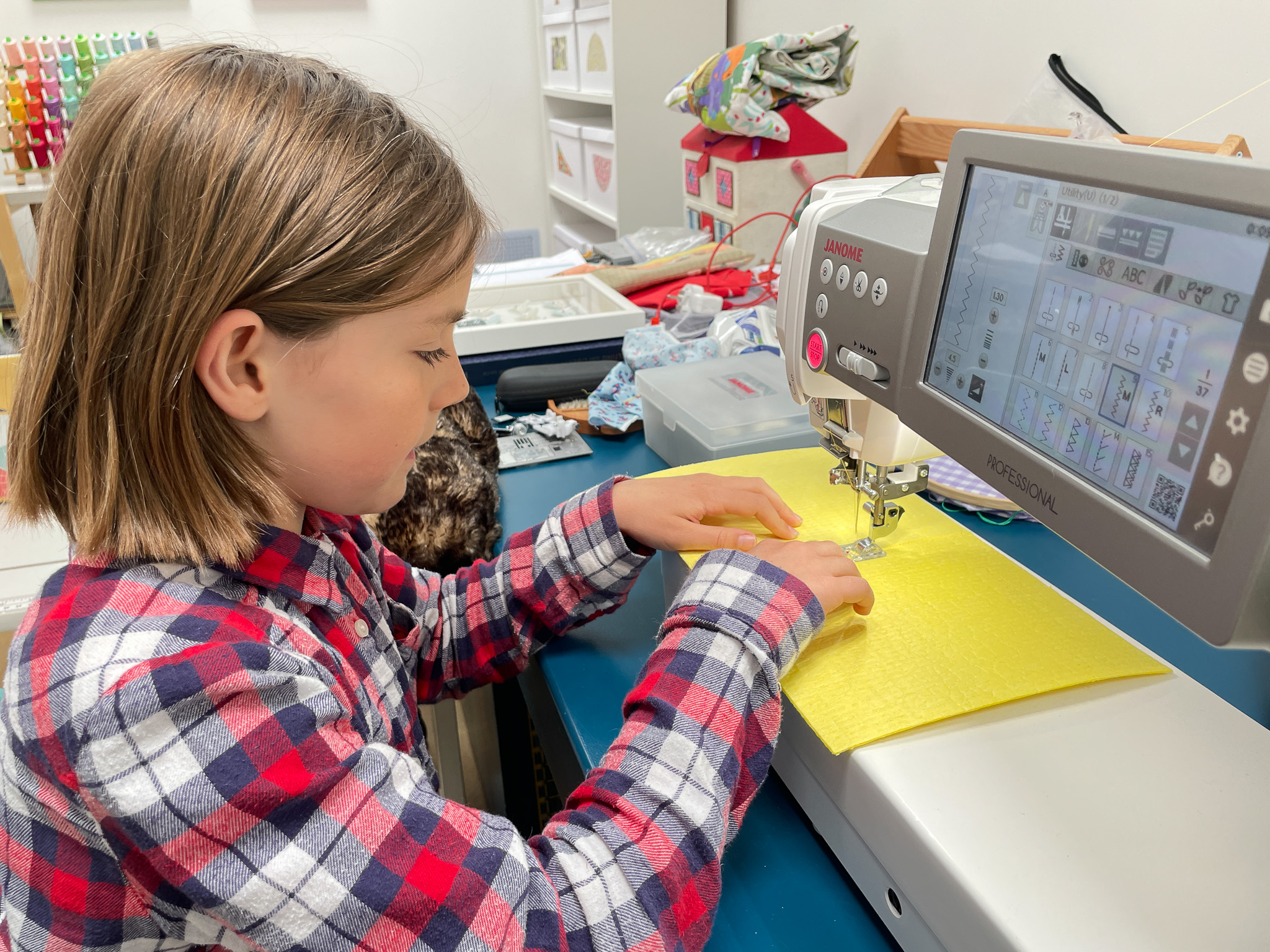 A young child in a red and blue checked shirt uses a sewing machine with yellow fabric in a bright, organised crafting room.