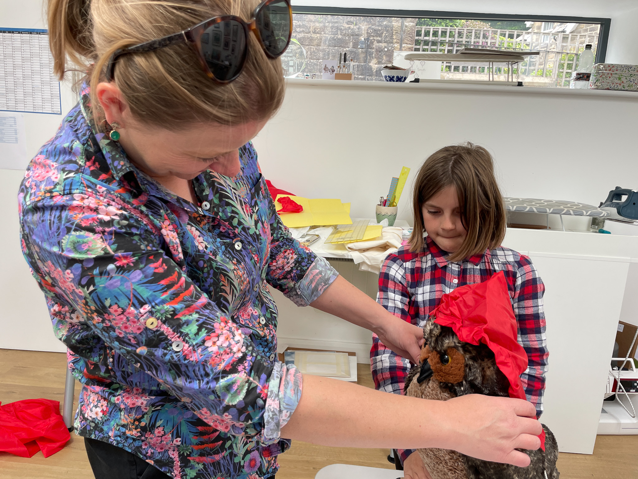 A woman helps a young girl put a red hat on a chicken indoors. The woman wears a floral blouse and sunglasses on her head; the girl wears a checked shirt and smiles at the chicken.