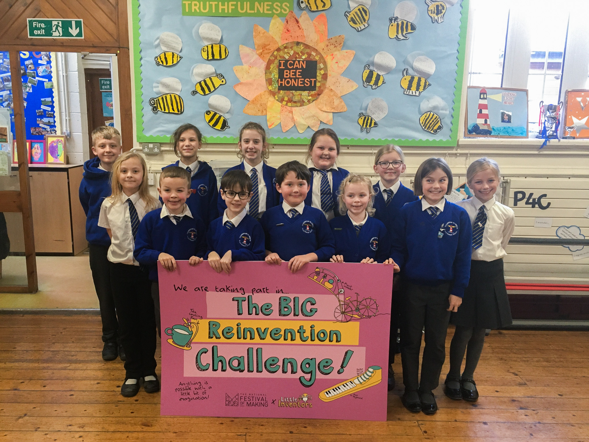 A group of school children in blue uniforms stand indoors, smiling and holding a colourful sign that reads The BIG Reinvention Challenge! The background features a decorated noticeboard with bees and inspirational words.