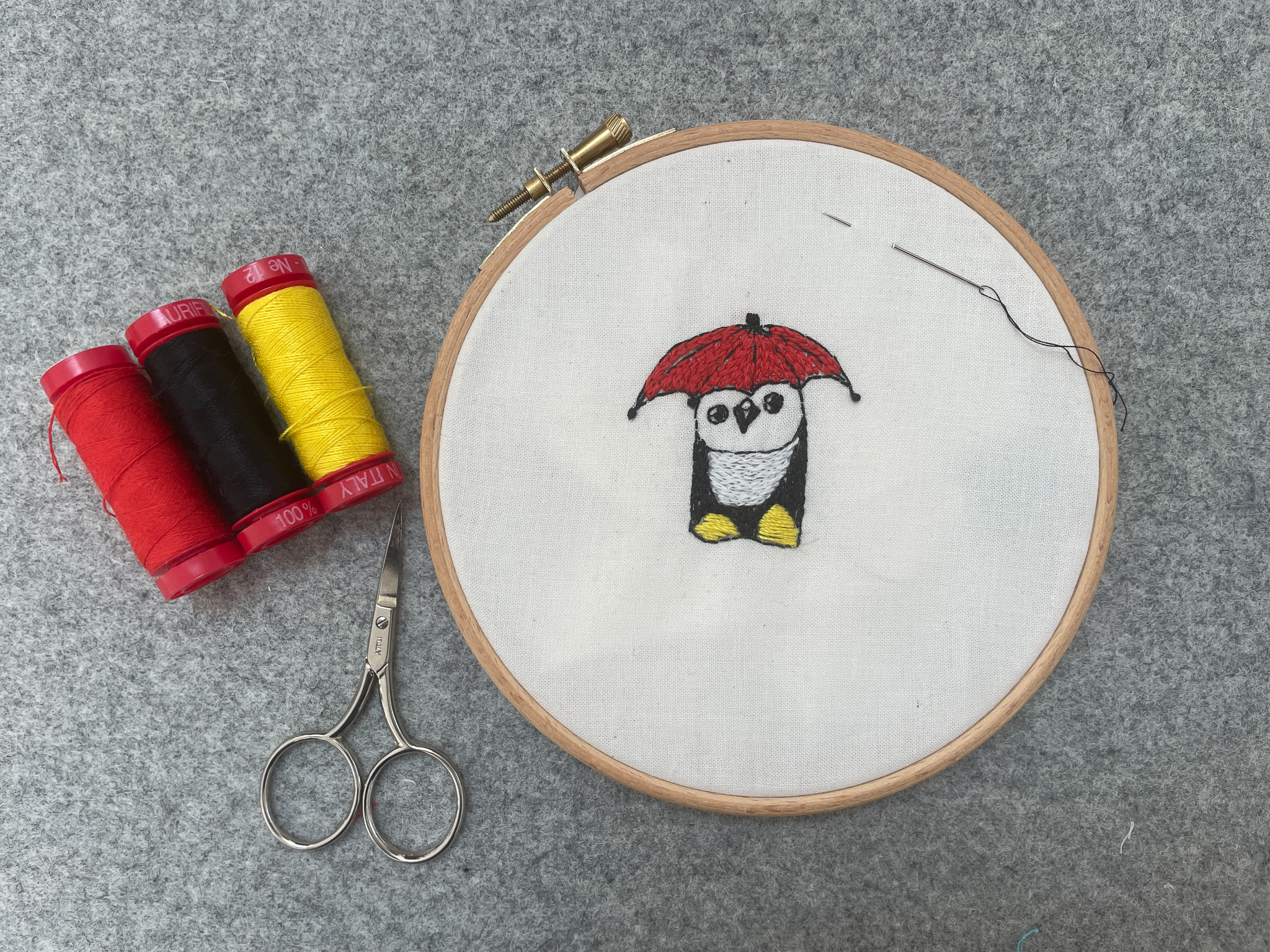 An embroidery hoop displays a stitched owl holding a red umbrella on white fabric. Nearby are red, yellow, and black thread reels and a pair of embroidery scissors, all on a grey surface.