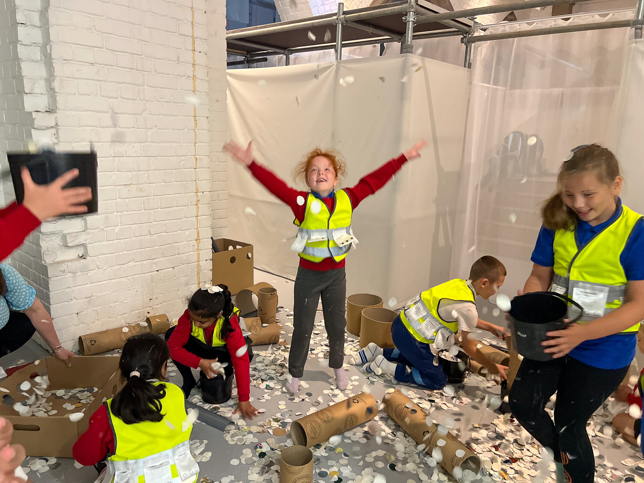 Several children wearing yellow safety vests play joyfully indoors, tossing white paper circles in the air. One child in the centre stands with arms stretched up, smiling excitedly, surrounded by playful chaos and cardboard tubes.