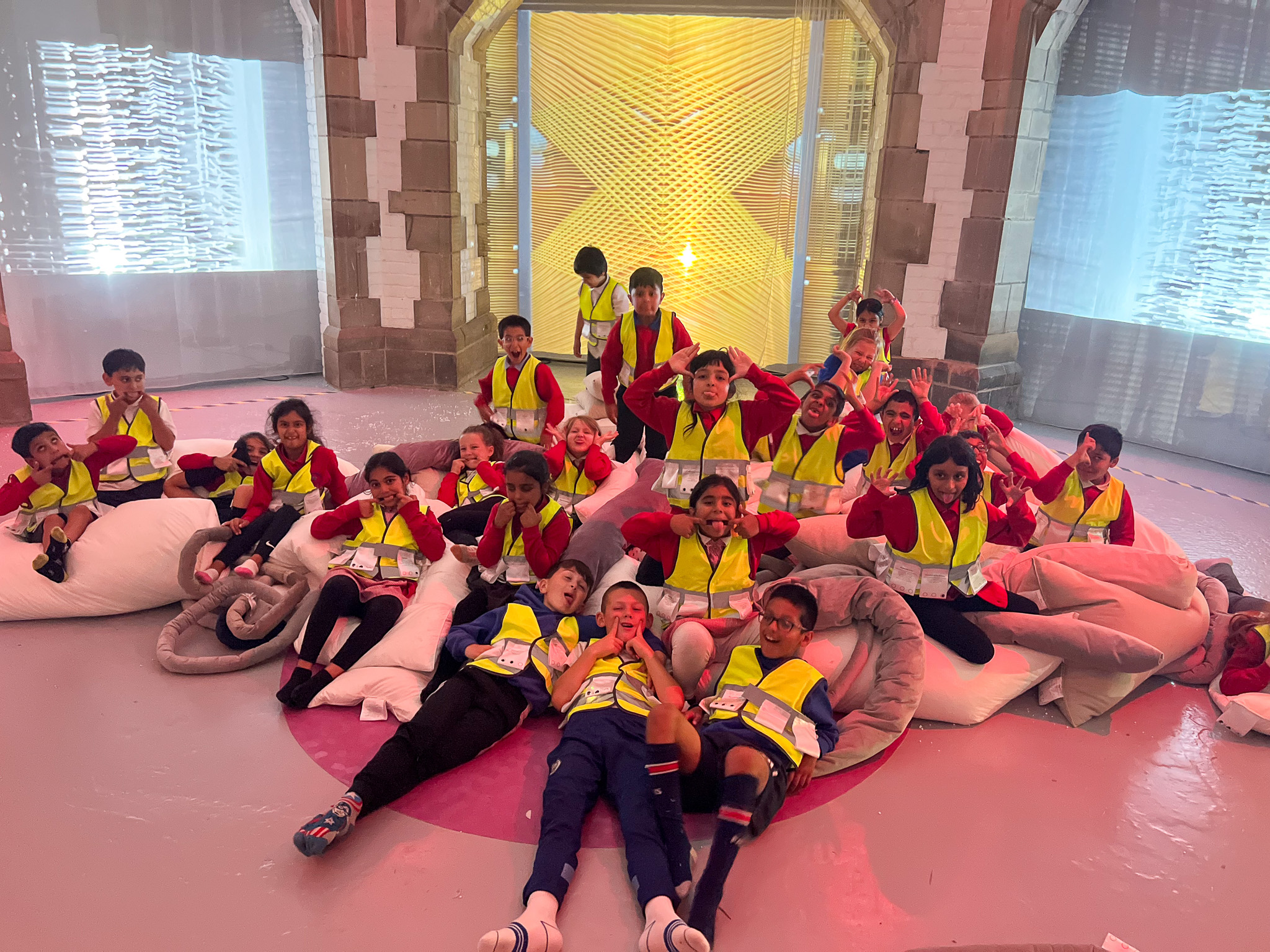 A group of children wearing yellow safety vests sit and lie on large, soft cushions in a brightly lit, playful indoor space, smiling and posing for a group photo.