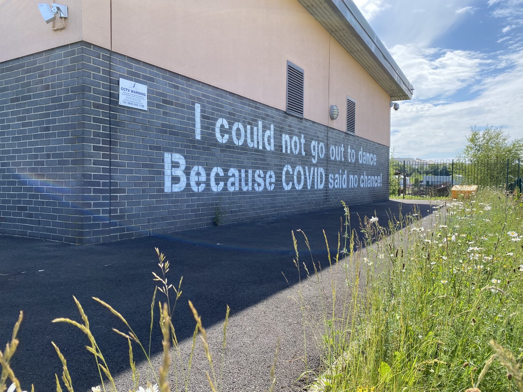 Sunny outdoor scene with a brick building, a wall reading “I could not go out to dance because COVID said no chance!”, and wildflowers lining the path.