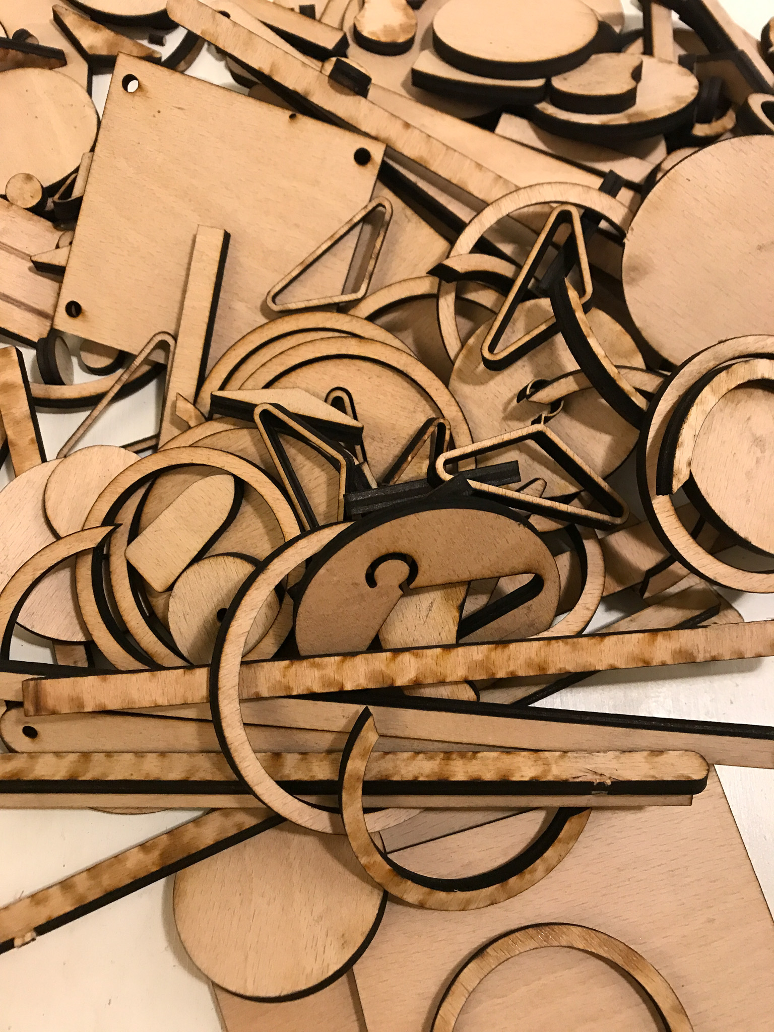 A pile of assorted wooden shapes, including circles, triangles, squares, and rectangles, with dark edges likely from laser cutting, scattered on a light surface.