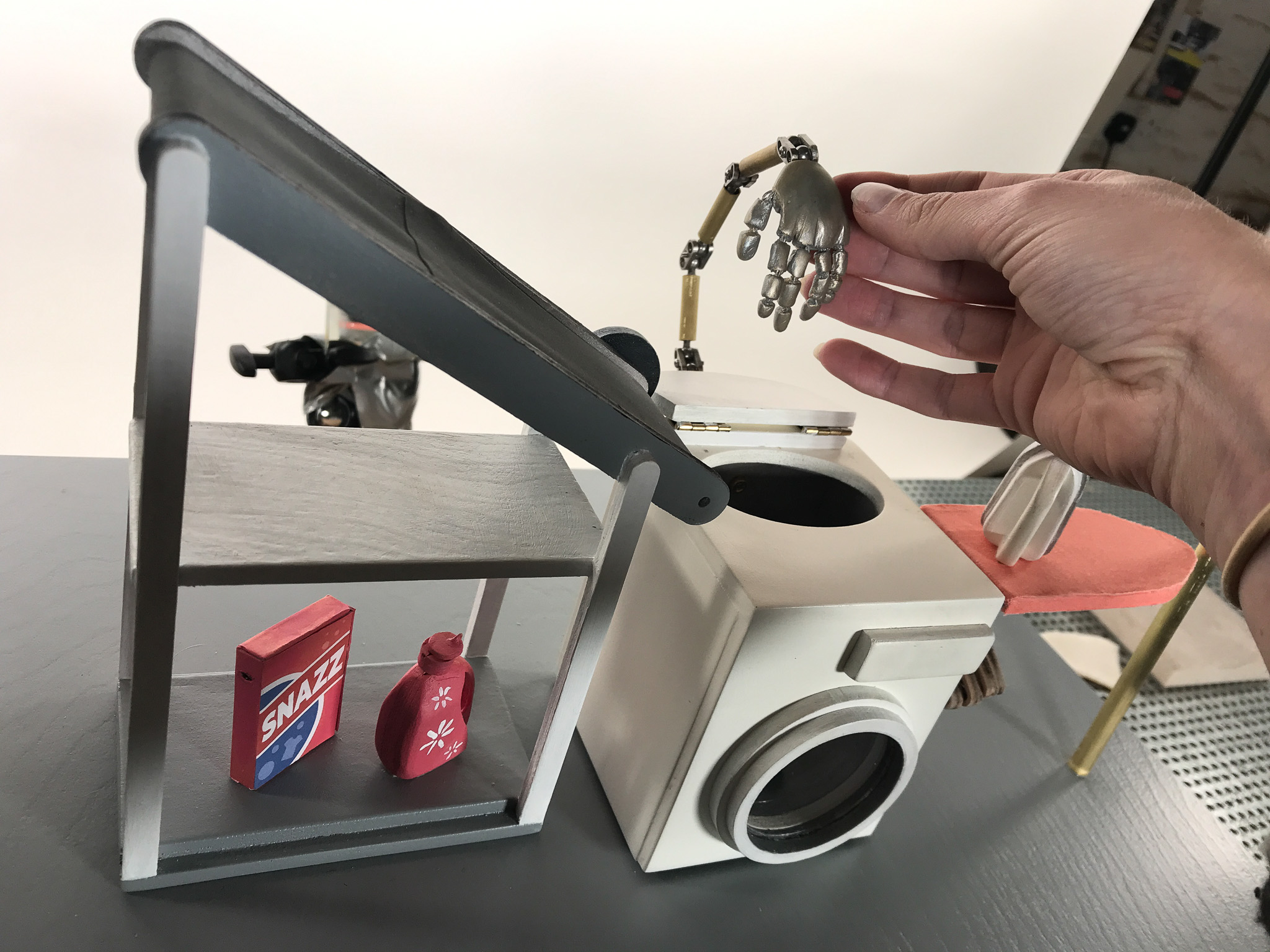 A hand holds a small articulated robot arm above a model of a washing machine. Nearby, a shelf displays a box labeled 