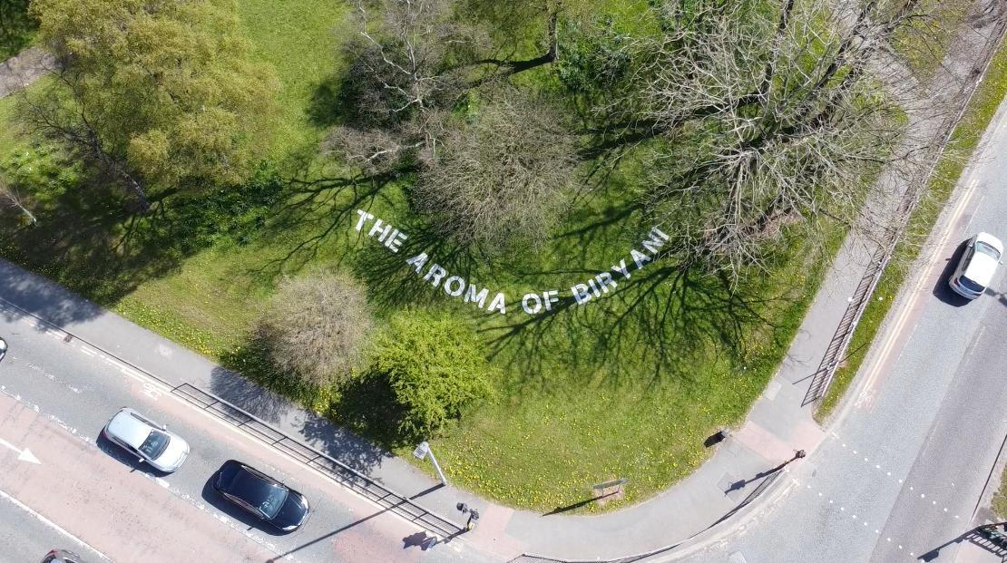 Aerial view of a grassy area with white letters on the ground spelling THE AROMA OF BIRNAM, surrounded by trees and next to a road with several cars.