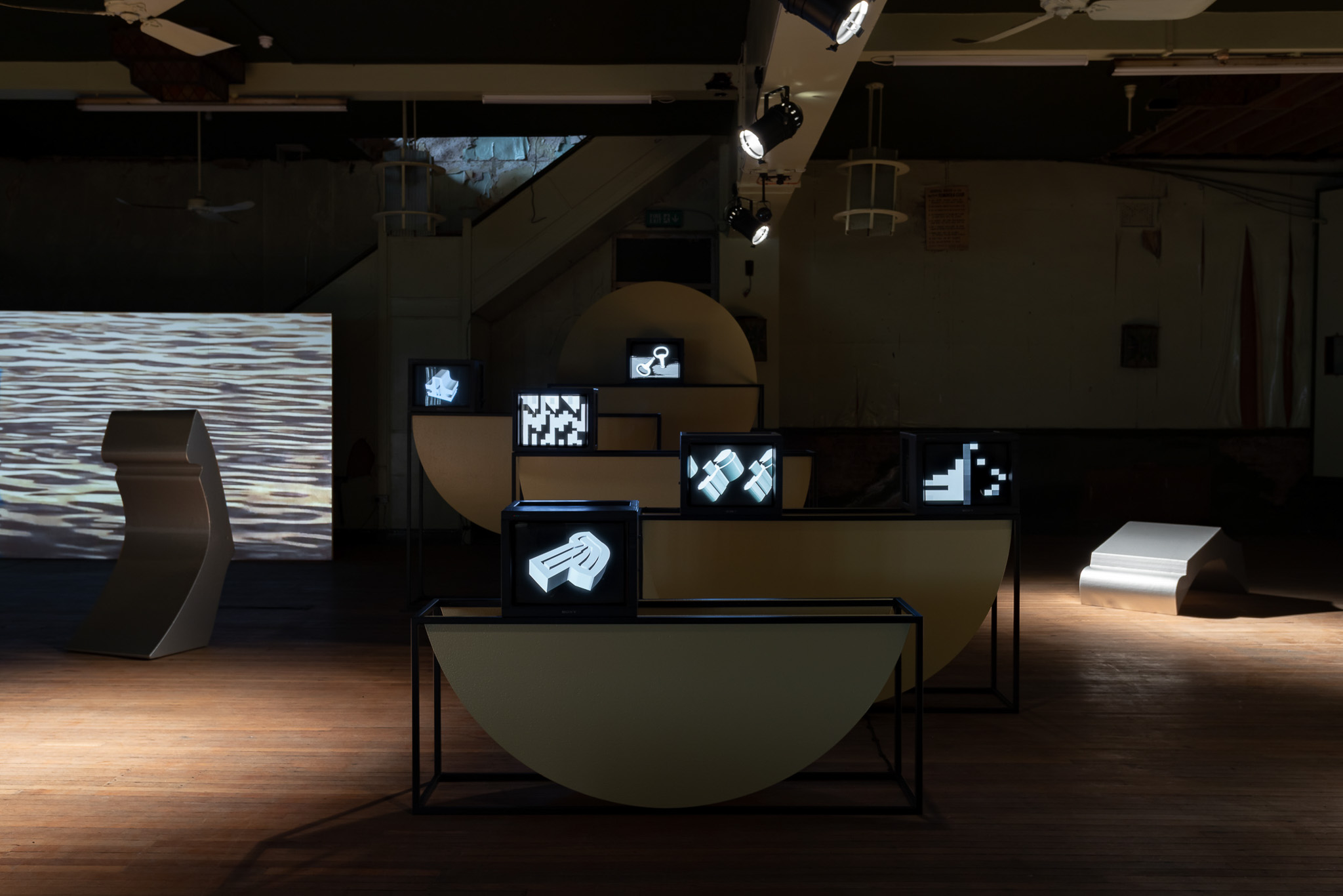 A dimly lit space displays geometric sculptures and screens showing black-and-white abstract digital images, arranged on curved and rectangular plinths on a wooden floor. Soft spotlights illuminate parts of the installation, and in the background, a large screen shows rippling water.