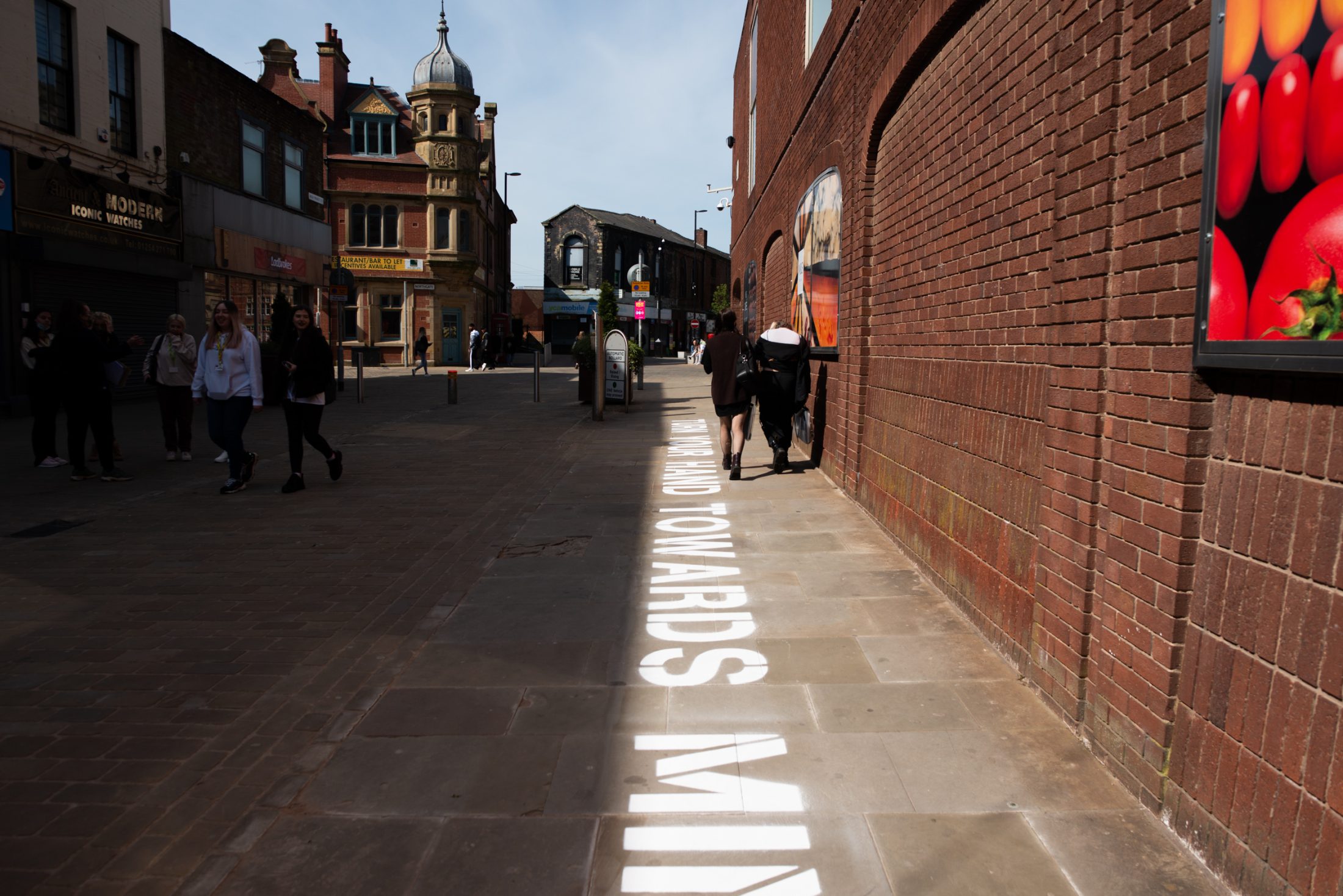 A sunlit street with people walking along a pavement next to a brick wall. Words painted on the road read AND TOWARDS MIN. Buildings and shops are visible in the background.