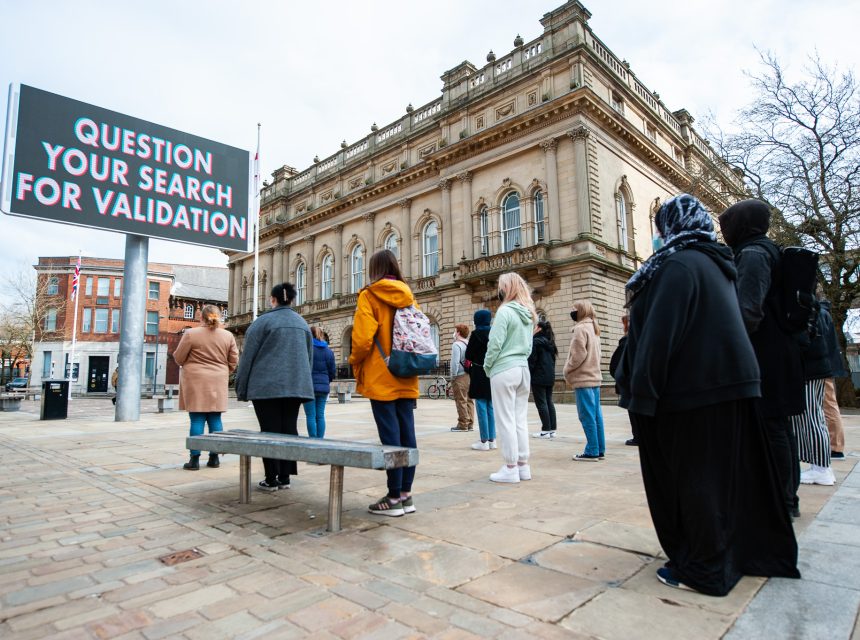 A group of people stand in front of a large building, looking at a hoarding that reads, QUESTION YOUR SEARCH FOR VALIDATION. The weather is clear and the setting is an urban square.