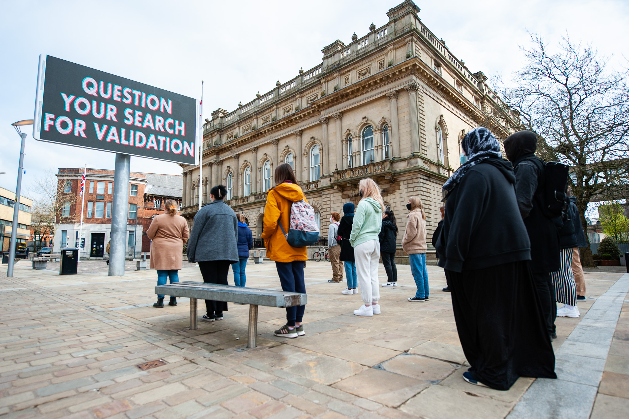 A group of people stand in front of a large building, looking at a hoarding that reads, QUESTION YOUR SEARCH FOR VALIDATION. The weather is clear and the setting is an urban square.