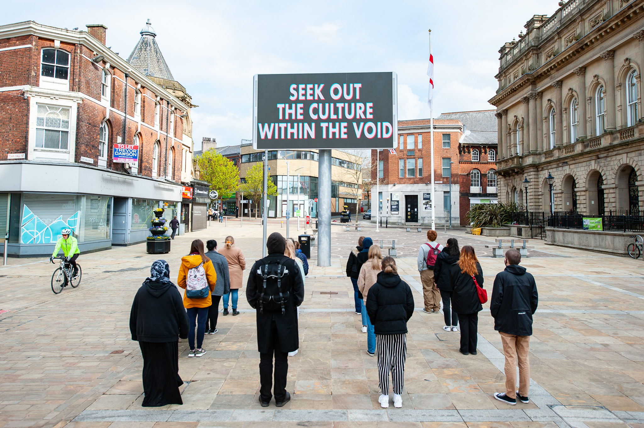 A group of people stands in a town square facing a large electronic sign that reads, SEEK OUT THE CULTURE WITHIN THE VOID. The surrounding buildings are historic and the area looks quiet.