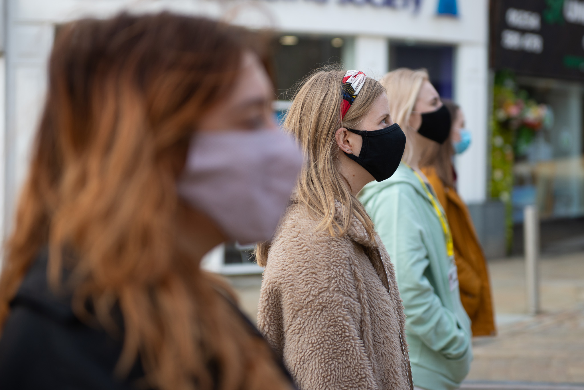 Three young women wearing face masks stand outdoors in a row, looking ahead. The background is urban and slightly blurred, suggesting a city street or shopping area.