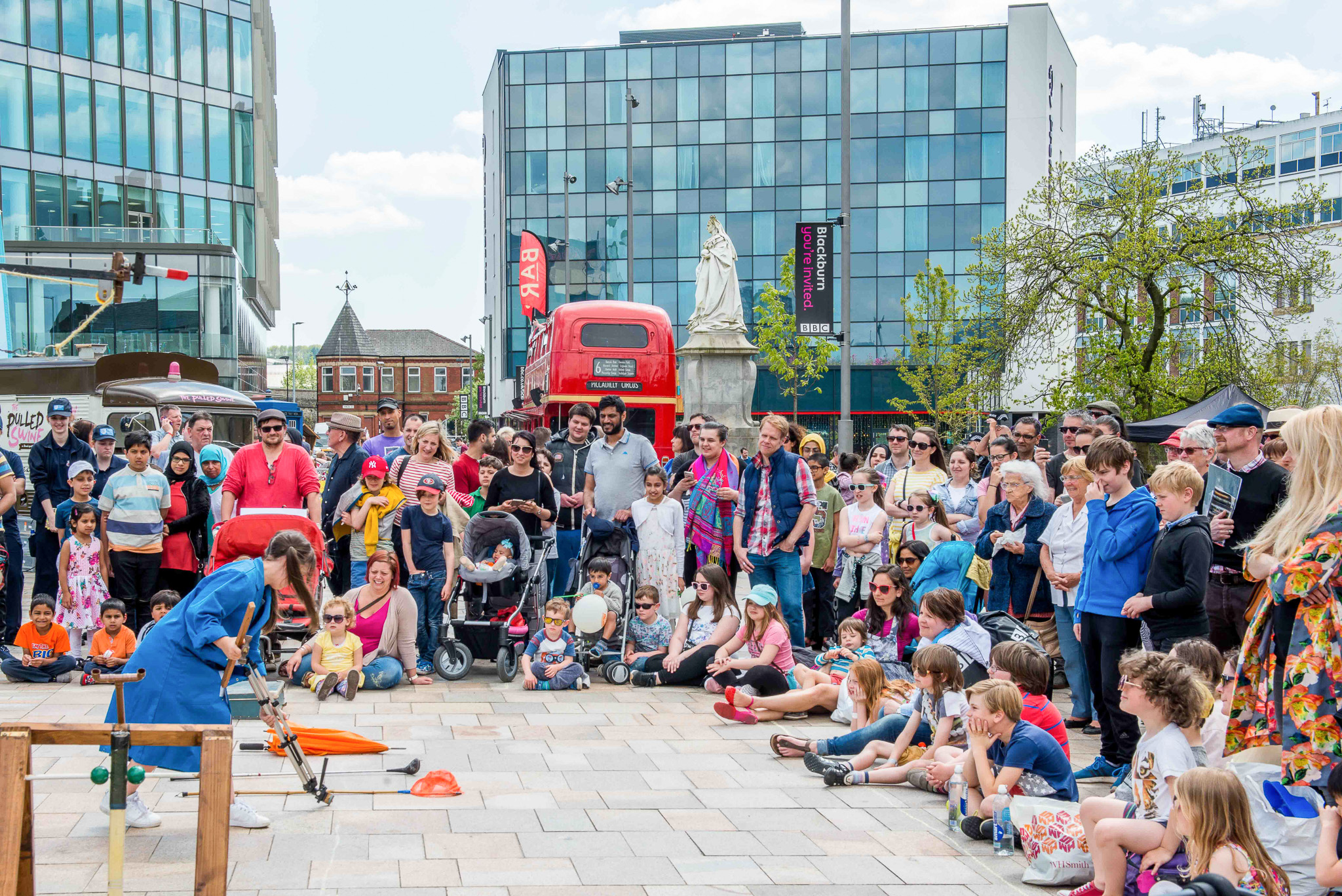 A street performer entertains a large, diverse crowd in a city square. People of all ages watch attentively, some seated on the ground, surrounded by modern buildings, a red bus, and a statue in the background.