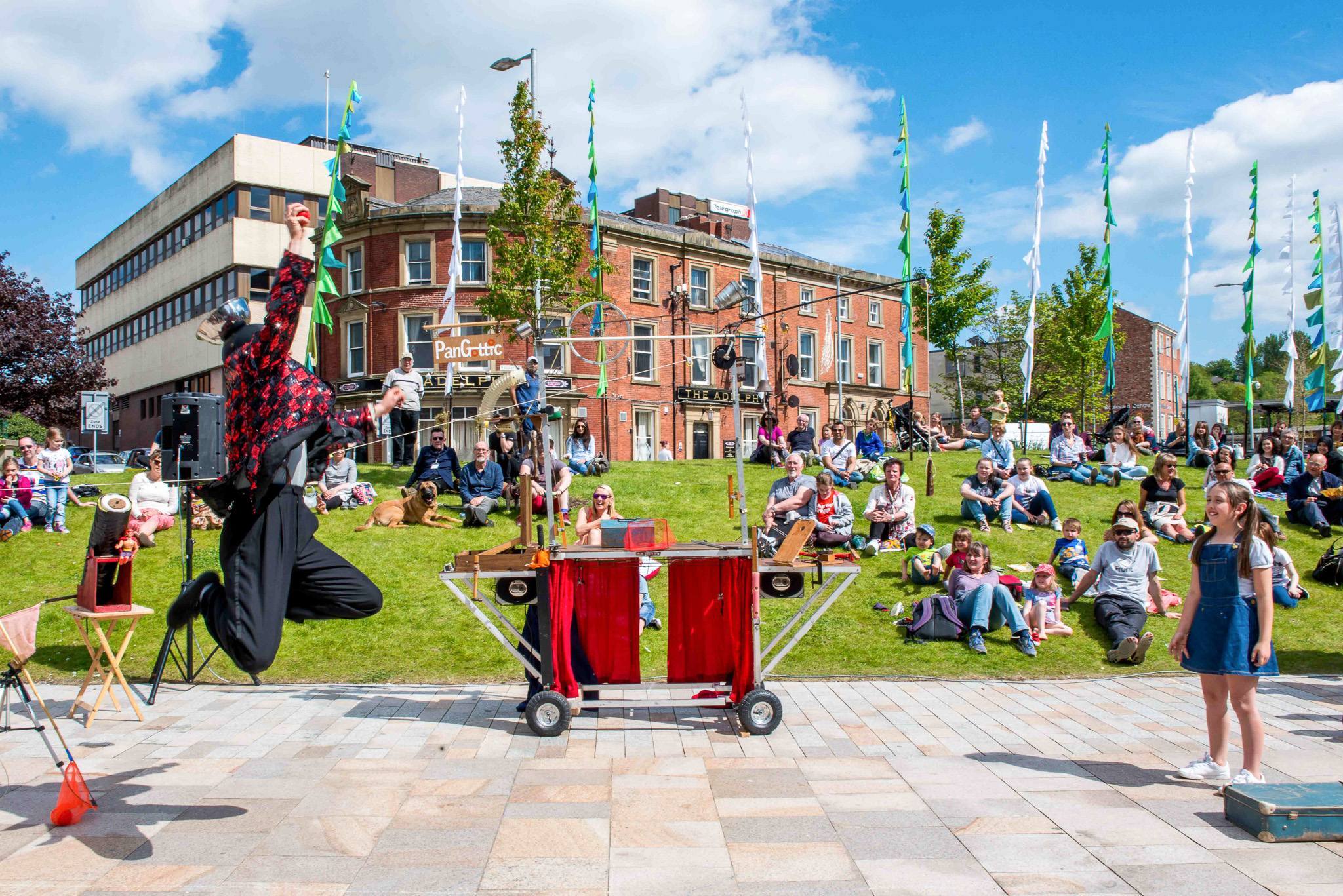 A street performer in colourful clothes jumps in the air during a show in front of an audience seated on grass. Props are arranged on a trolley, and a young girl stands nearby. Buildings and trees are in the background under a blue sky.