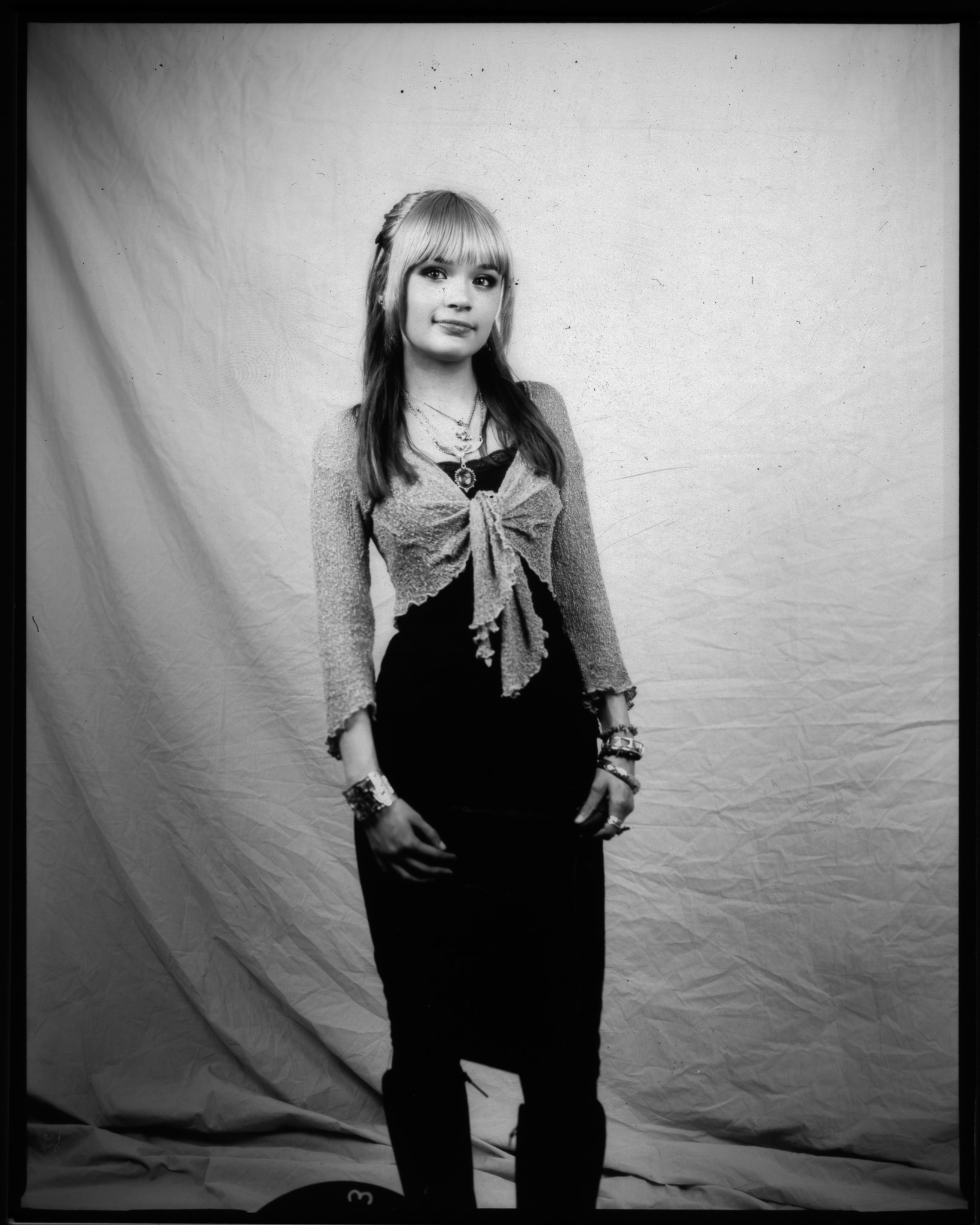 A person standing against a plain backdrop, wearing a tied top and black skirt, with accessories and long hair.