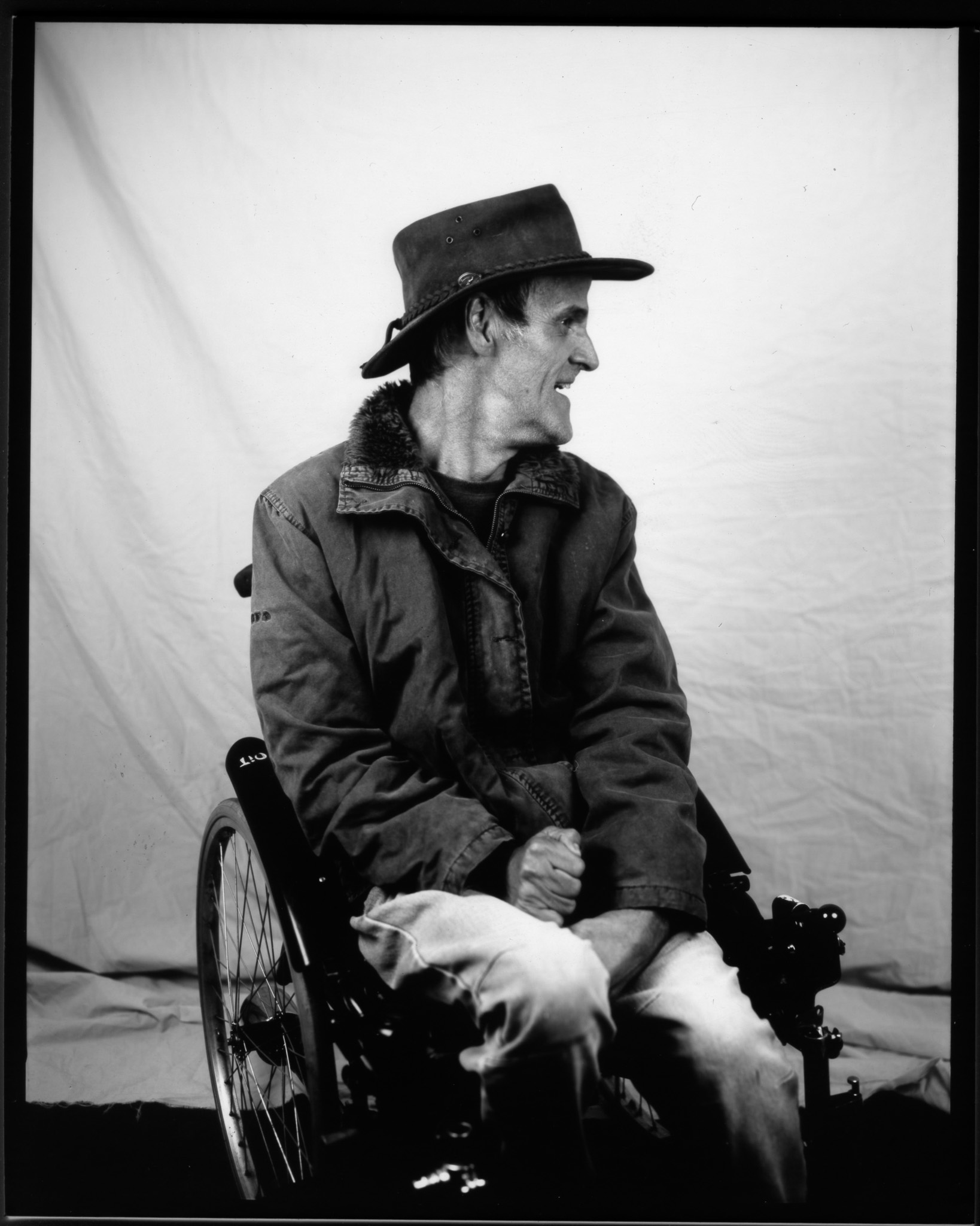 A man in a wheelchair wearing a hat and a jacket, sitting sideways against a plain background.