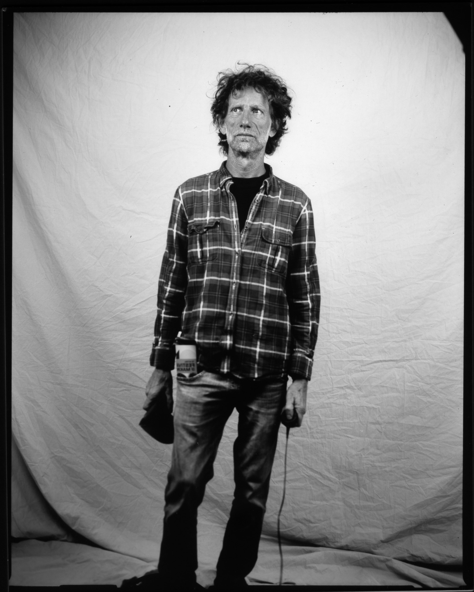 A person standing against a plain background, wearing a flannel shirt and jeans, with tousled hair.