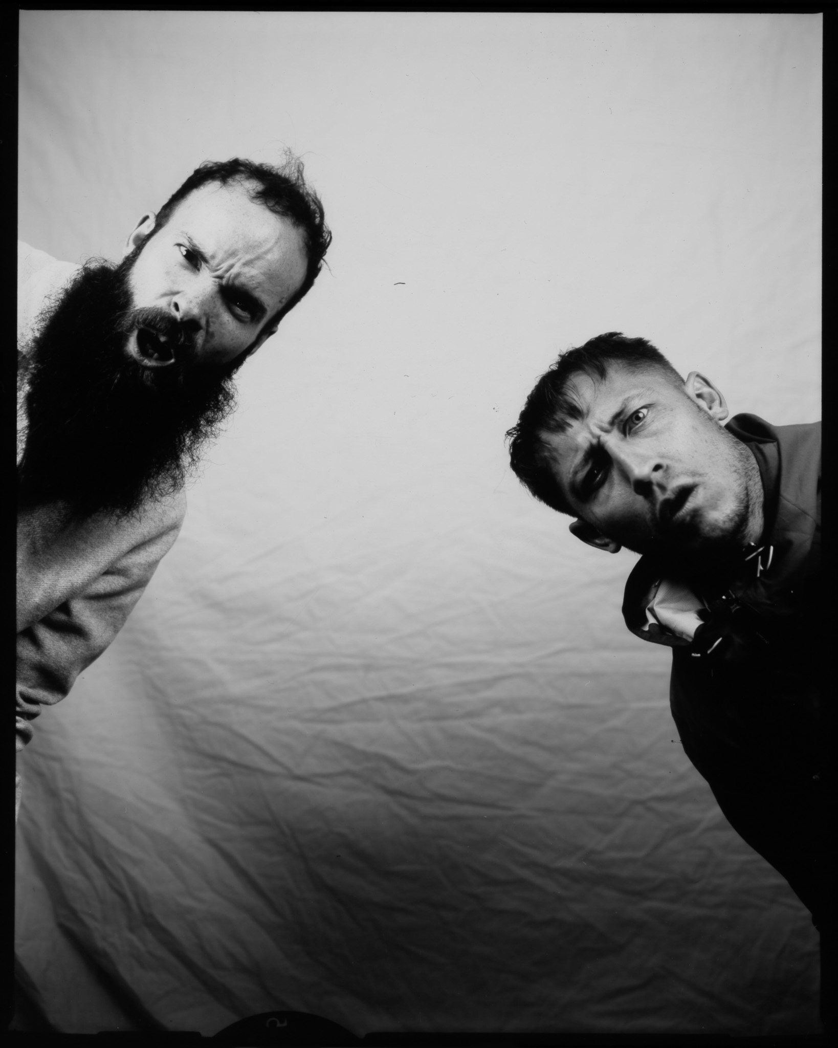 Two men with expressions of surprise, leaning towards the camera against a plain background. One has a long beard and the other has shorter hair.