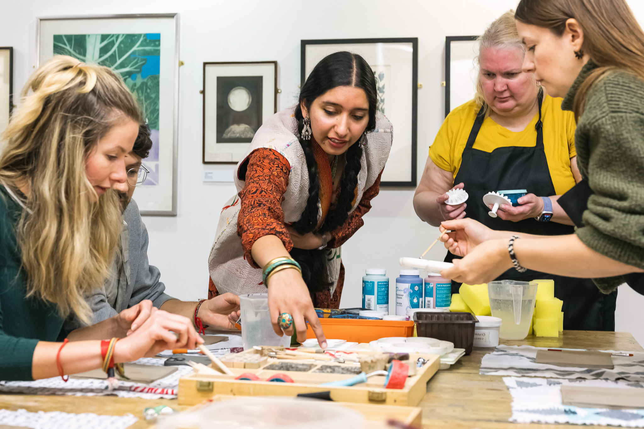 A group of women sitting and standing around a table, engaged in an art or craft activity with paint and tools. One woman in the centre points at a project, while others work with brushes and materials. Artworks hang on the wall behind them.
