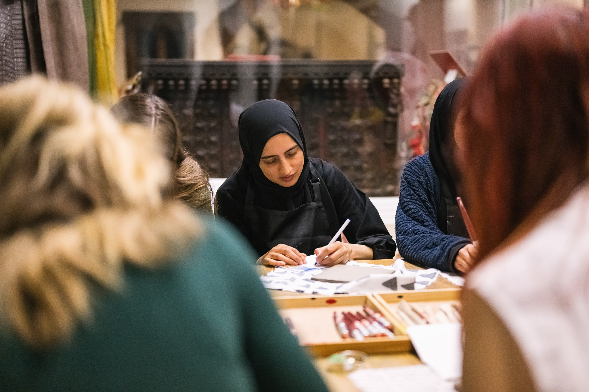 A woman wearing a black hijab and apron is seated at a table, concentrating on writing or drawing on paper, surrounded by others who are also engaged in creative activities.