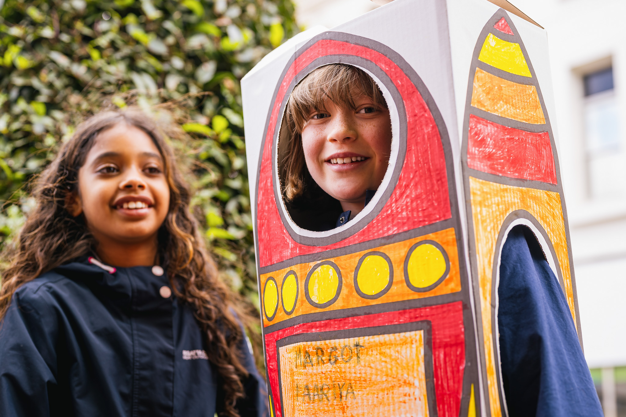 Two children smiling, one wearing a colourful cardboard rocket costume with a green leafy background.