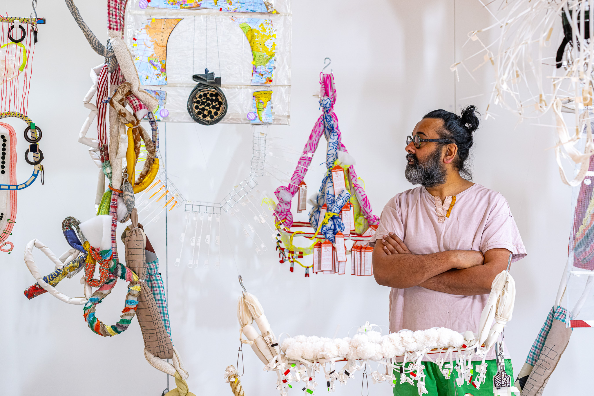 A man with a beard and glasses stands with his arms crossed, looking at colourful, eclectic textile and mixed-media art pieces hanging on a white wall. The scene is vibrant and creative.
