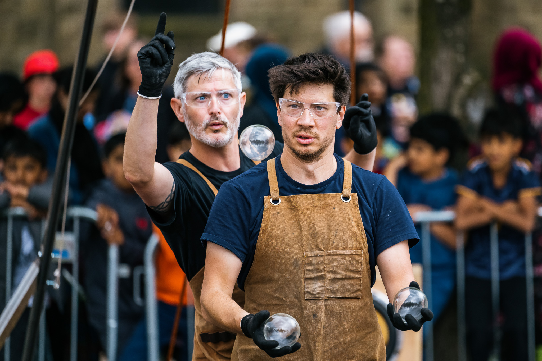 Two men wearing brown aprons, glasses, and black gloves perform with clear acrylic balls outdoors, surrounded by a crowd of onlookers behind barriers. One man points upwards while the other looks ahead, holding balls in both hands.