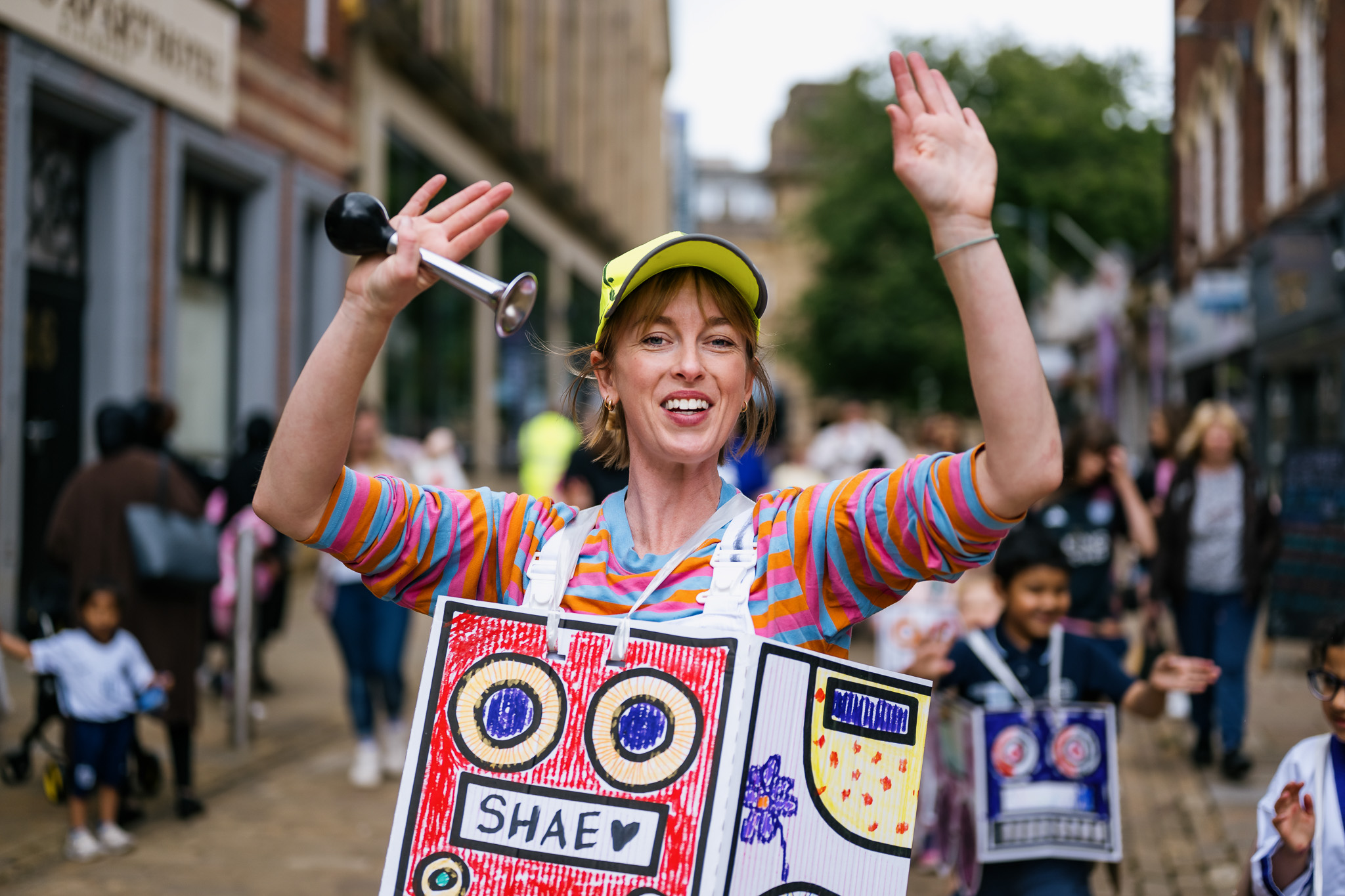A smiling person wearing a yellow cap and colourful striped shirt raises their hands at an outdoor event. They wear a decorated cardboard box with drawings and the name Shae on it. People walk in the background on a city street.