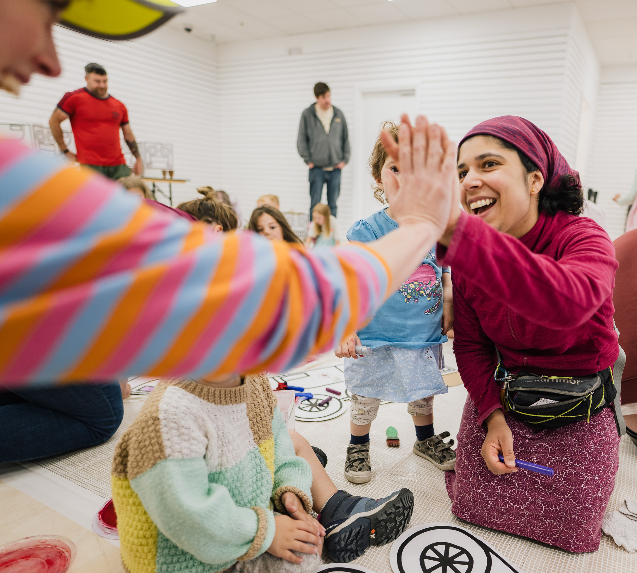 A smiling woman in a pink headscarf gives a high five to someone in a colourful striped jumper, surrounded by children and adults in a bright indoor space.