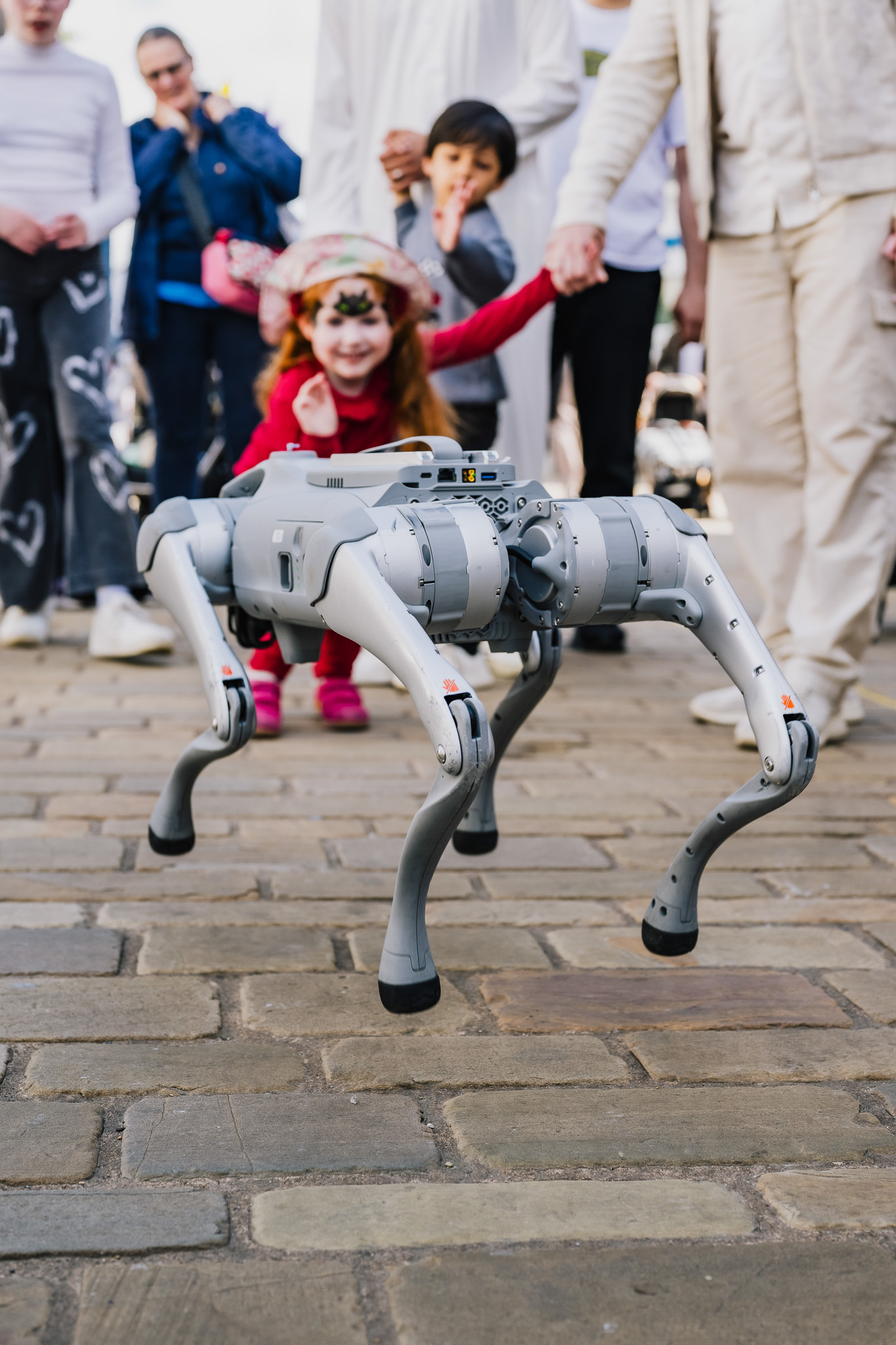 A robotic dog walks on a cobblestone path, captivating a smiling child in a red outfit and hat, surrounded by onlookers.