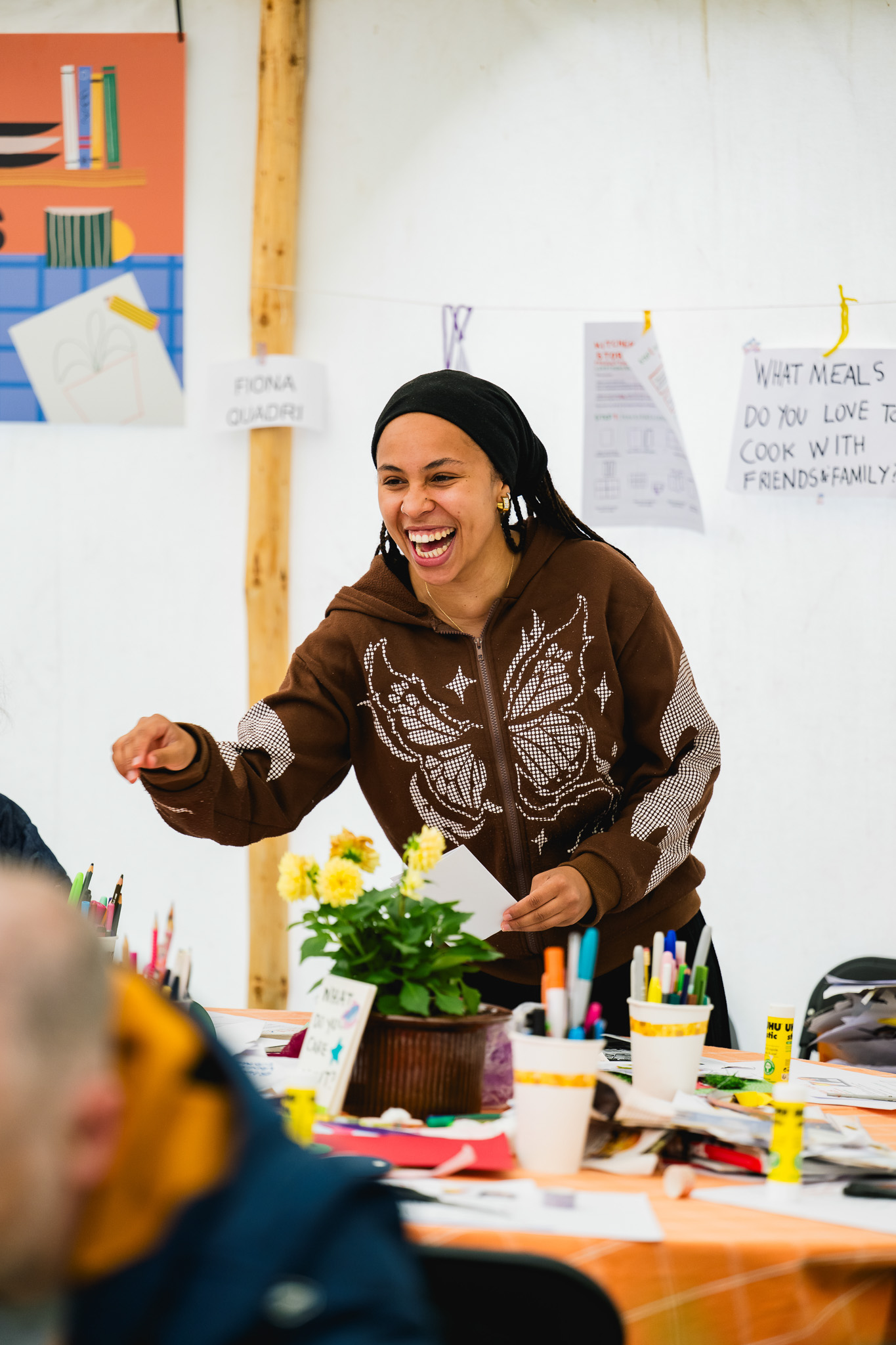 A woman smiles and gestures whilst standing at a table covered with art supplies and flowers. Papers, signs, and drawings are displayed behind her on a white wall. The scene appears lively and creative.
