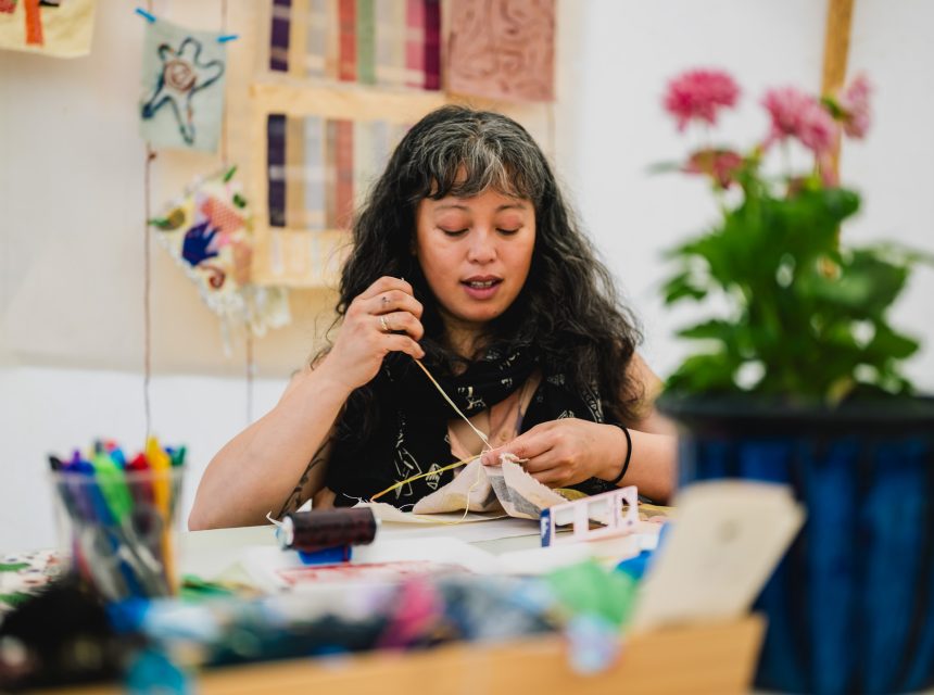 A woman with long, wavy hair sits at a table, focused on hand-stitching fabric. Colourful art supplies and flowers are on the table, and textile artworks hang on the wall behind her.