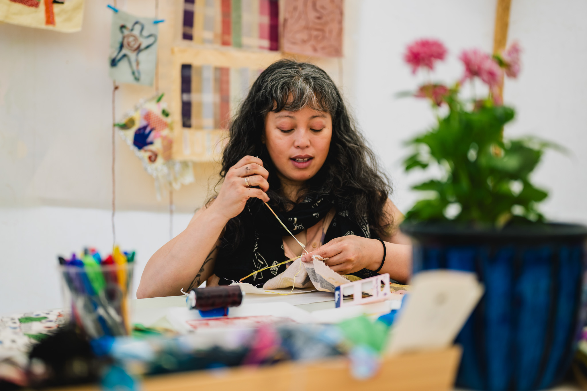 A woman with long, wavy hair sits at a table, focused on hand-stitching fabric. Colourful art supplies and flowers are on the table, and textile artworks hang on the wall behind her.