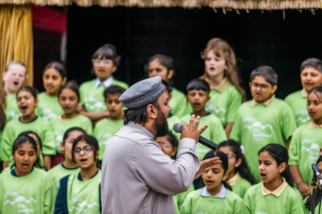 A man sings into a microphone on stage whilst a group of children in matching green shirts sing behind him.
