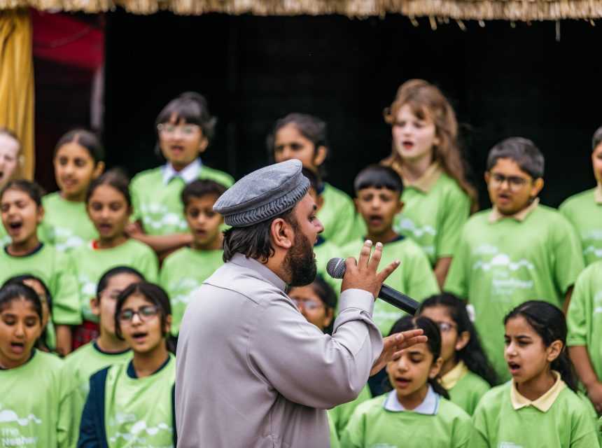 A man sings into a microphone on stage whilst a group of children in matching green shirts sing behind him.