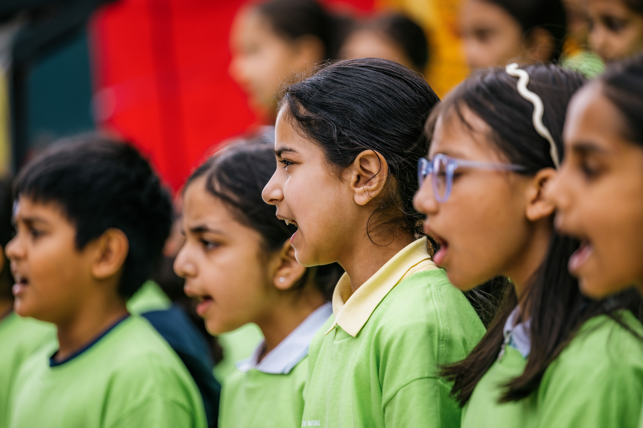 Children in green shirts sing in a choir, with blurred red and yellow colours in the background.