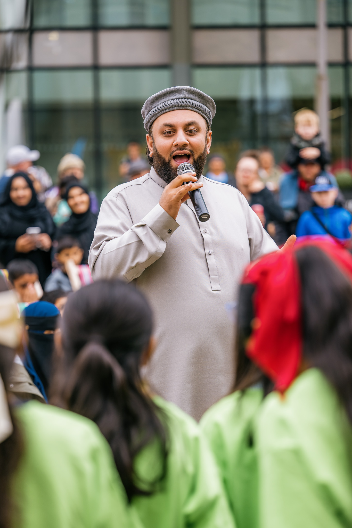 Man in traditional attire sings into a microphone at an outdoor event, facing children in green while the crowd listens attentively.