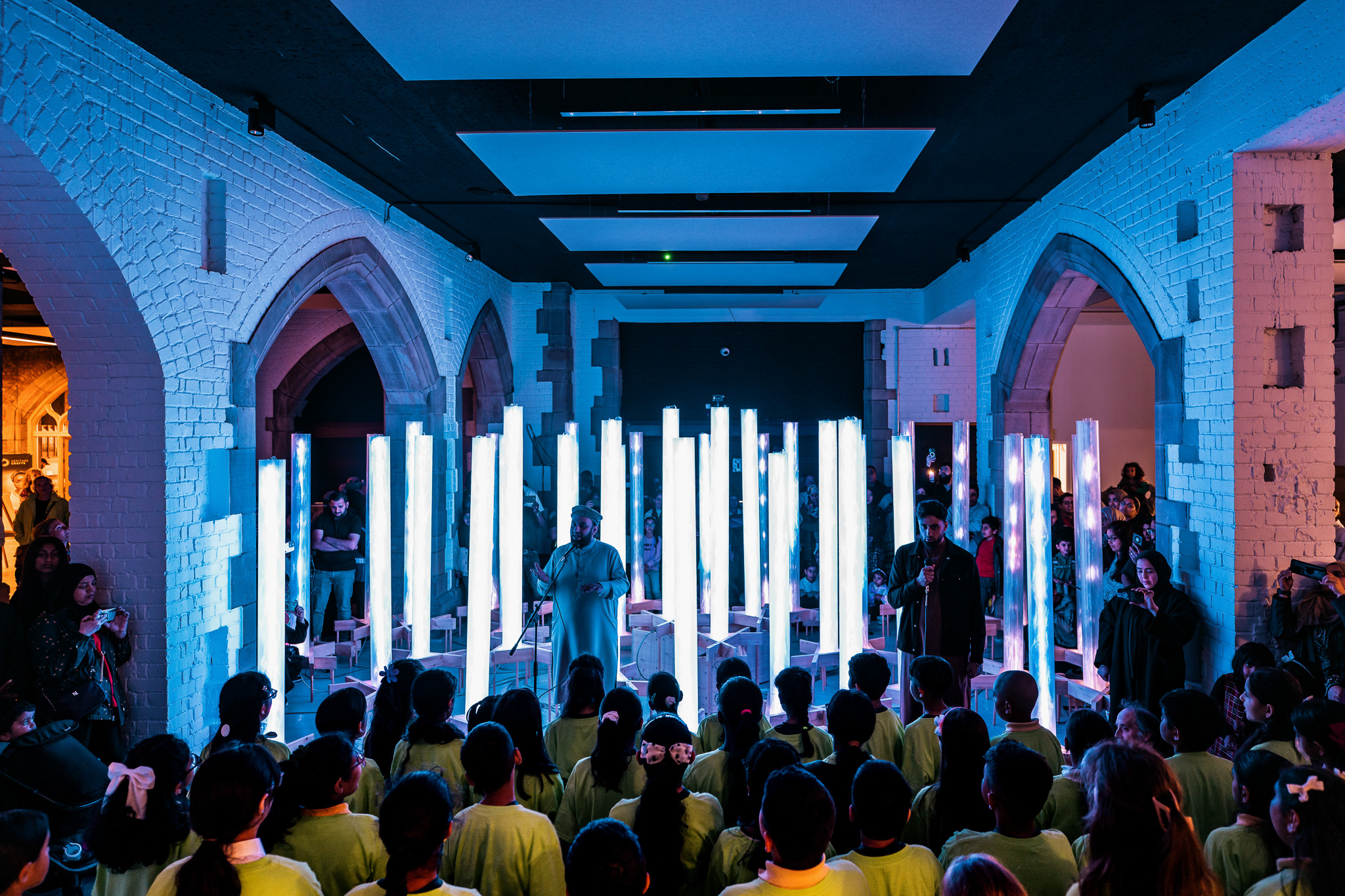 A group of children in yellow shirts watch a vibrant light installation featuring vertical, glowing pillars inside a large hall with arched brick walls and blue lighting. Adults stand near the exhibit, observing.