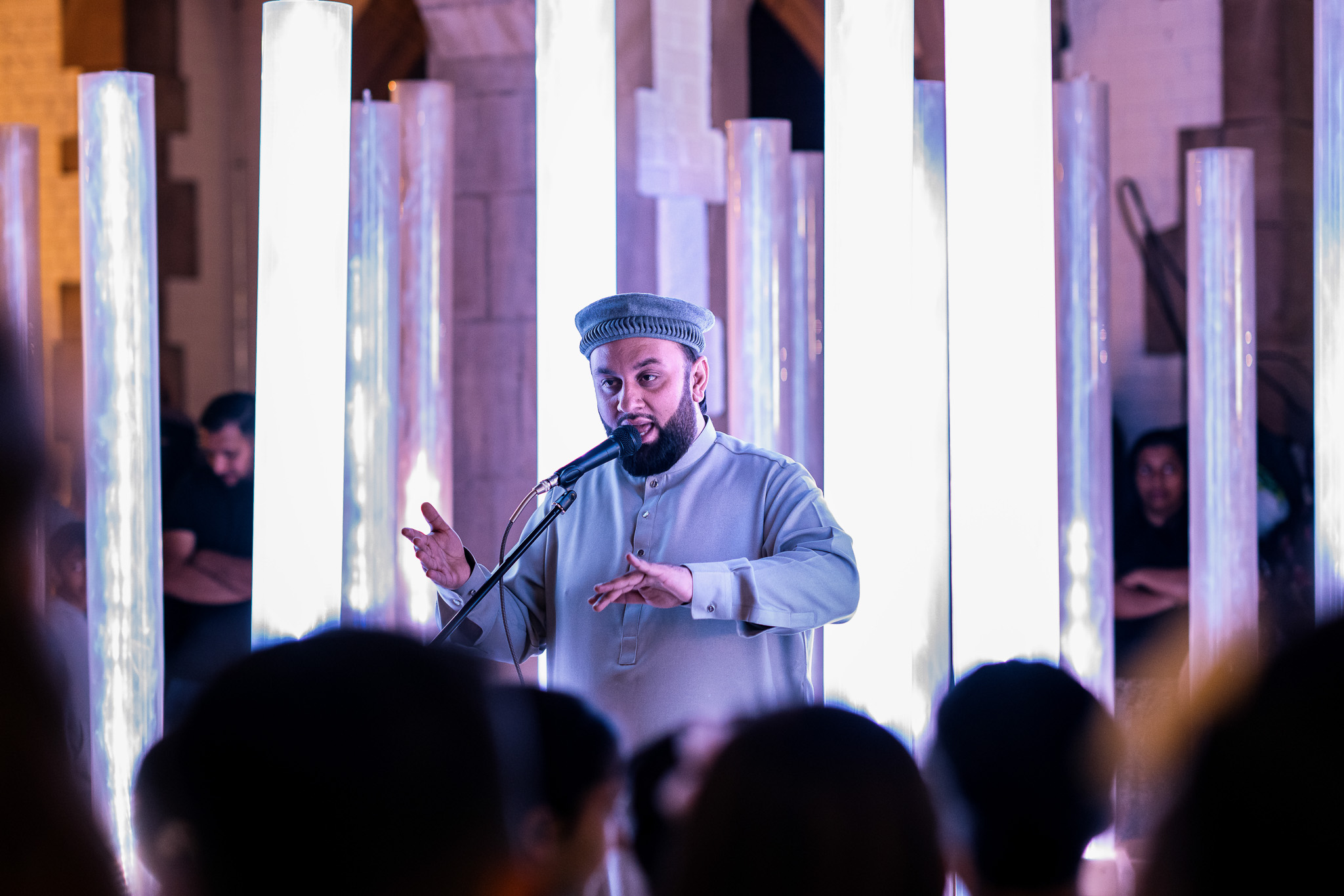 A man in traditional attire sings into a microphone on stage, surrounded by tall, glowing vertical light columns, with an audience watching in the foreground.