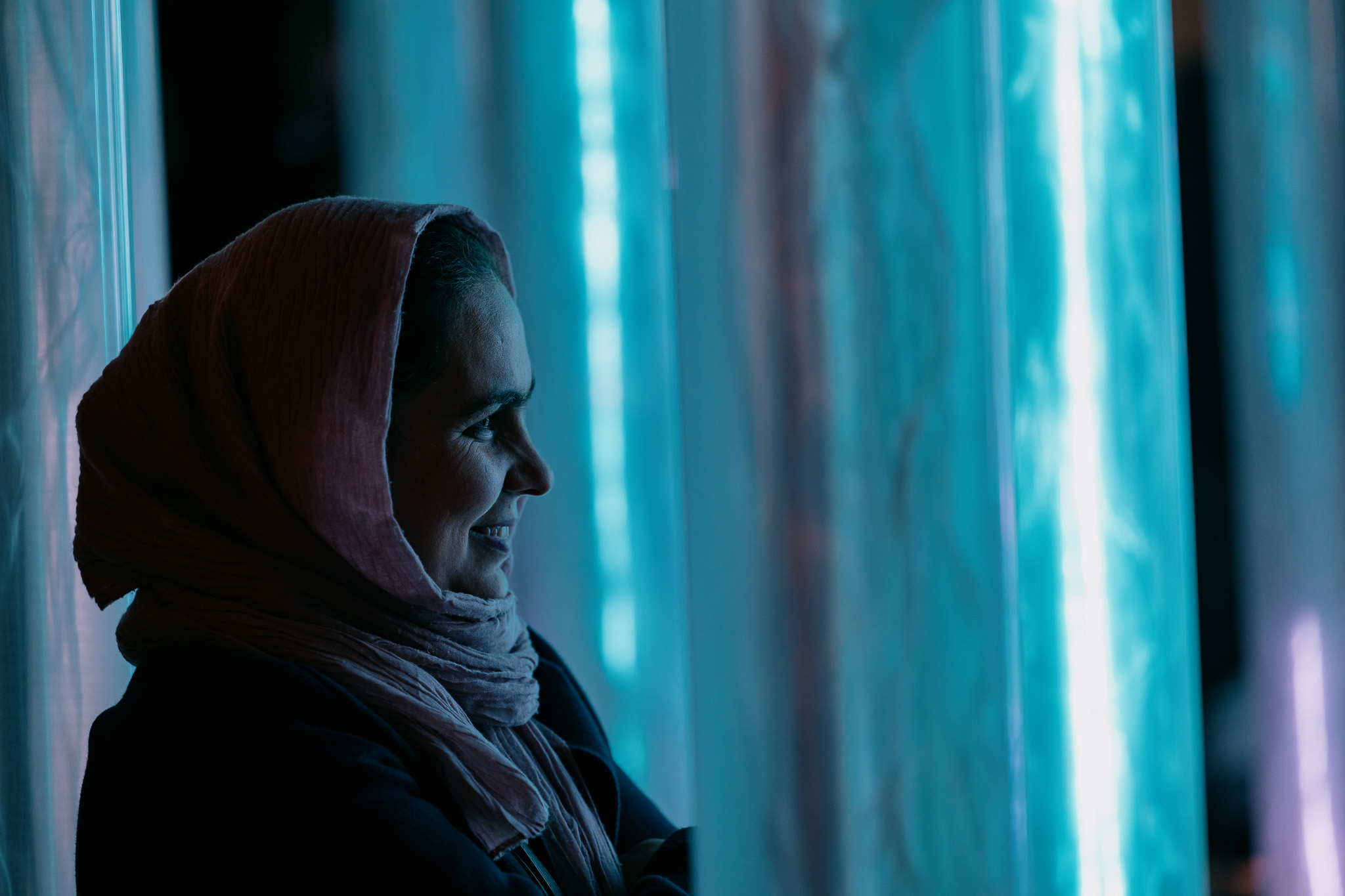 Woman in a hijab stands inside a translucent blue art installation, softly lit and smiling.