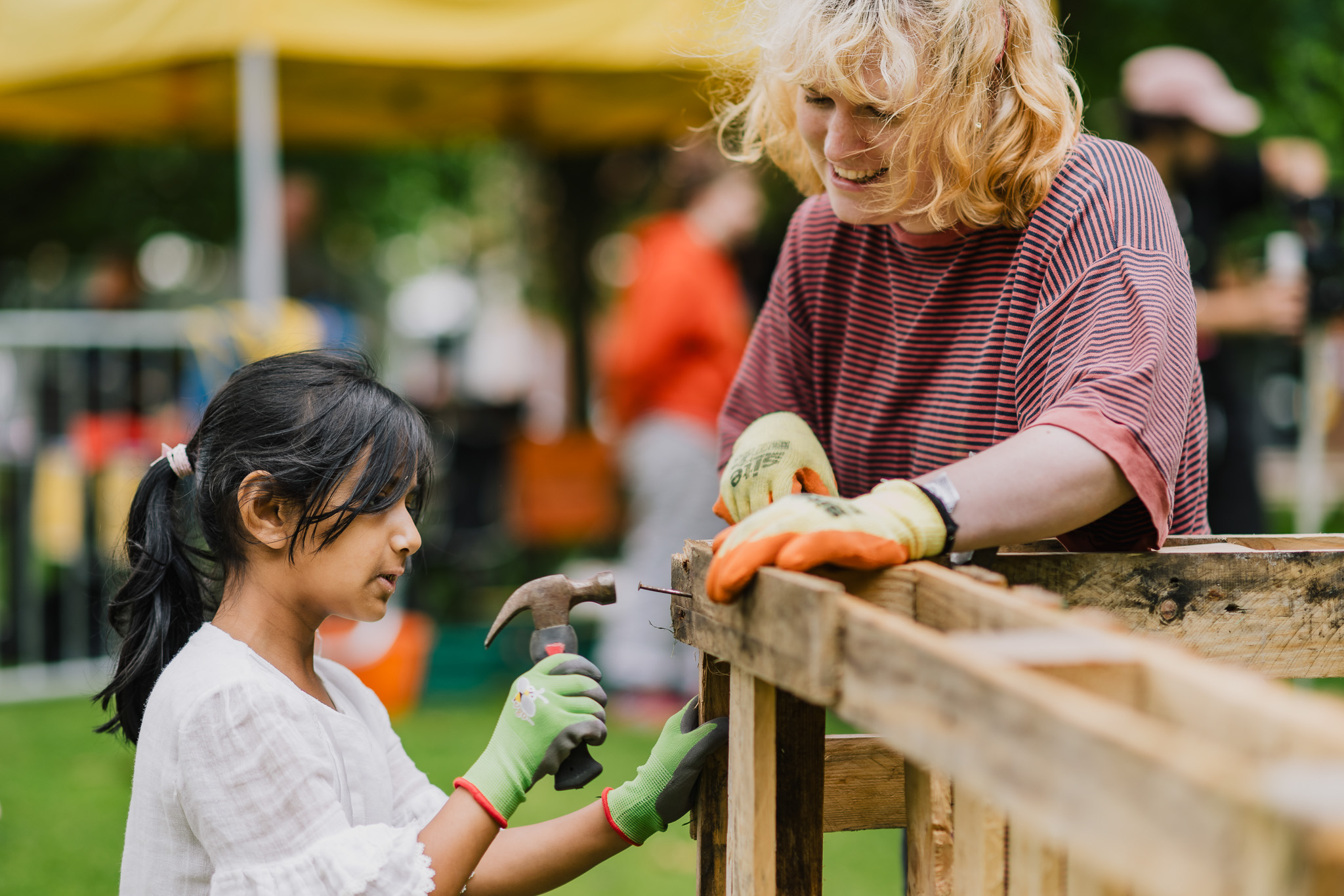 A young girl and an adult wearing gloves work together outdoors, building a wooden structure. The girl holds a hammer, and both are smiling. There is greenery and a yellow tent in the background.