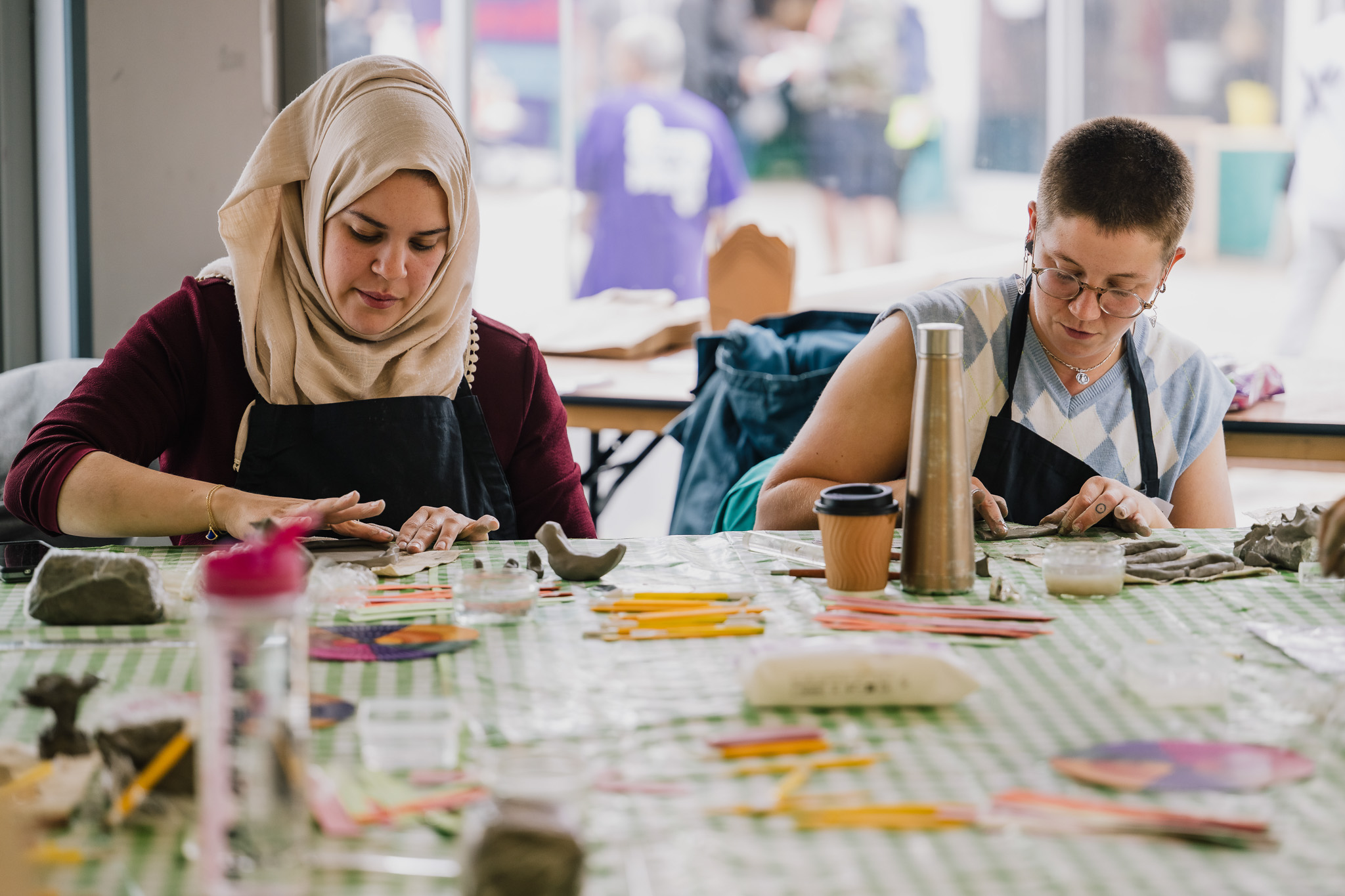 Two women sit at a table covered with art supplies and clay, focusing on their work. One wears a hijab and the other has short hair and glasses. The setting appears to be a creative workshop or art class.