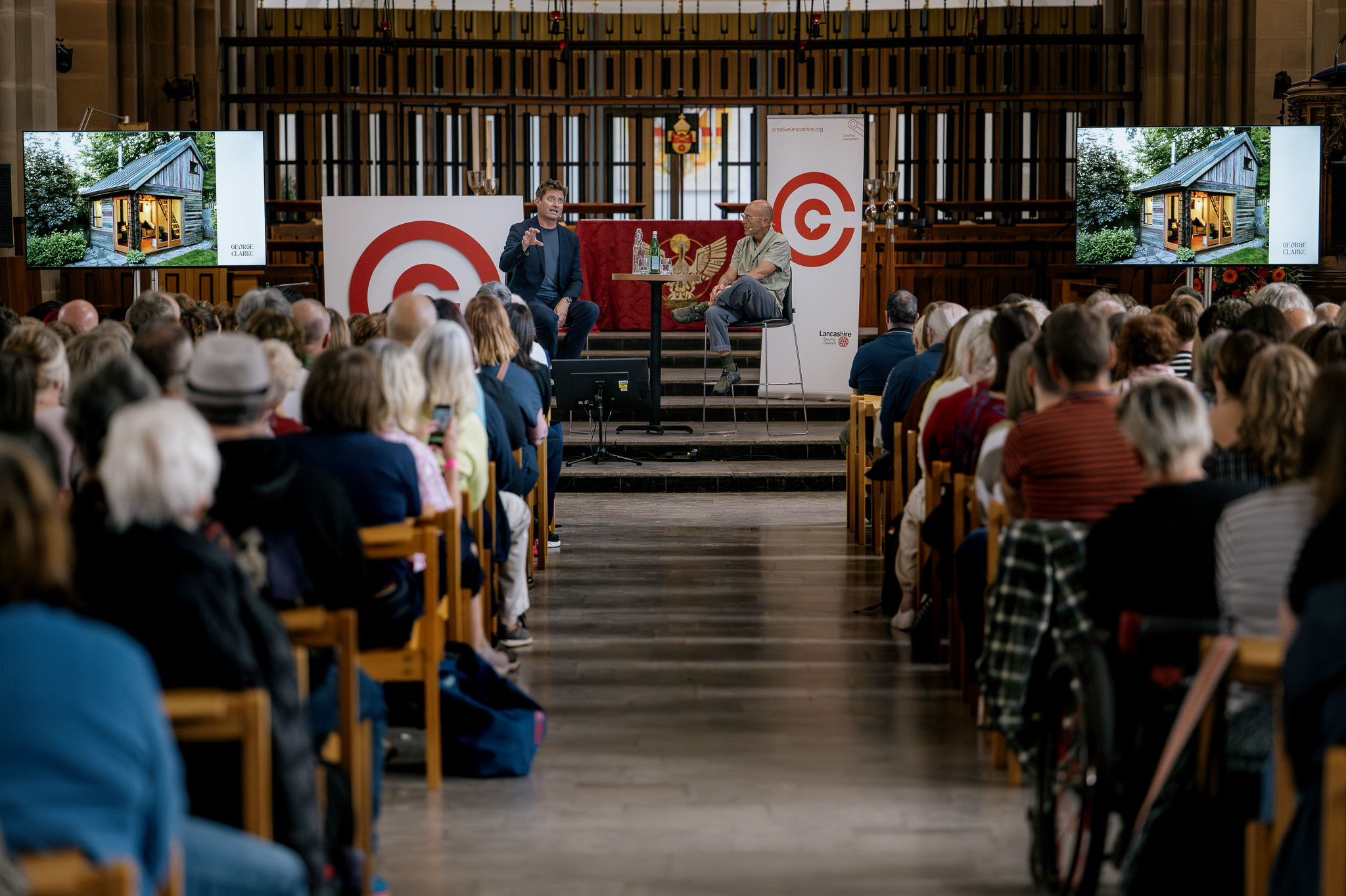 A large audience sits in rows facing two speakers, George Clarke and Wayne Hemmingway, on a stage in a cathedral. Two screens show an image of a wooden house, and banners behind the speakers.