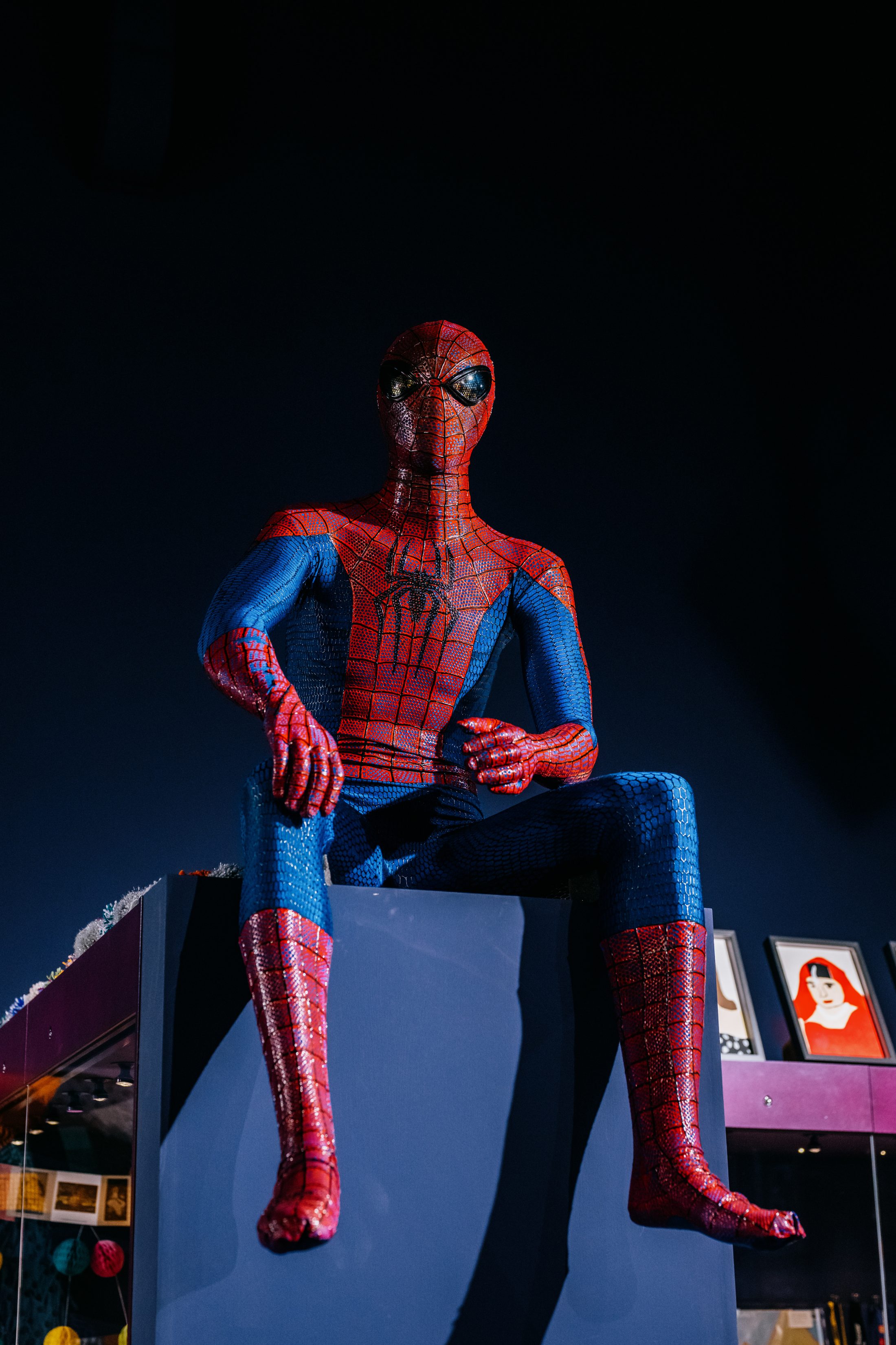 Spider-Man figure sits on a blue pedestal against a dark background, arms resting on knees.