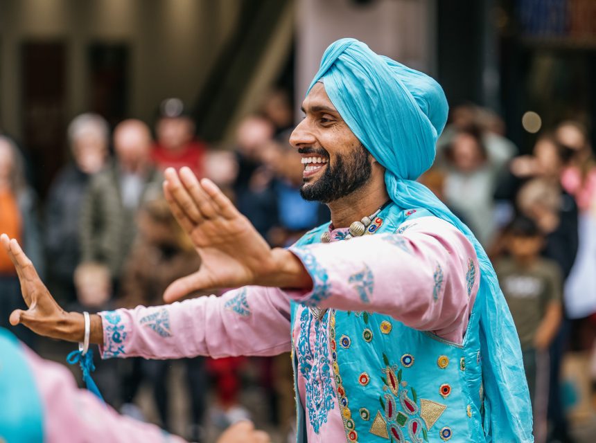 A man wearing a bright blue turban and embroidered outfit smiles and dances with his arms outstretched, performing at an outdoor event with a blurred crowd in the background.